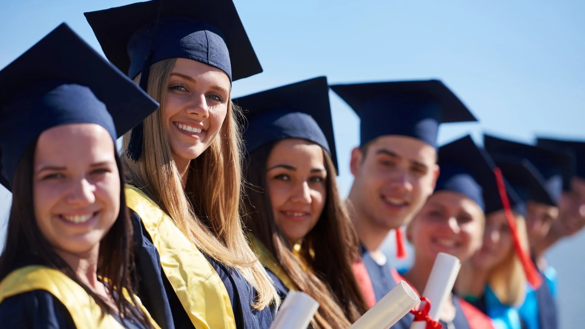 A group of graduates in caps and gowns smile and hold diplomas, lined up outdoors under a bright blue sky.
