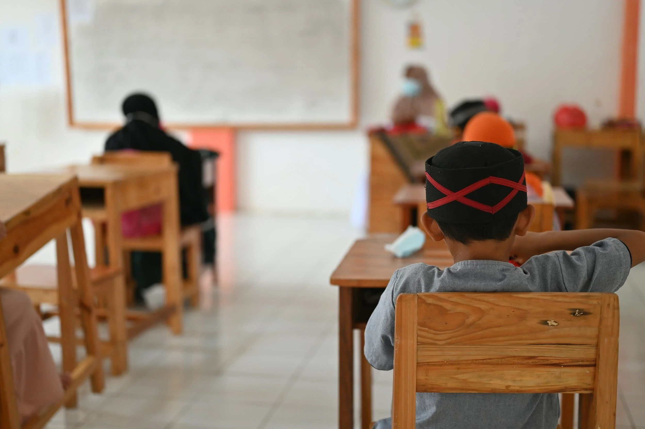 A young boy sits at a wooden desk in a classroom, facing the front, with other students seated at desks ahead of him.