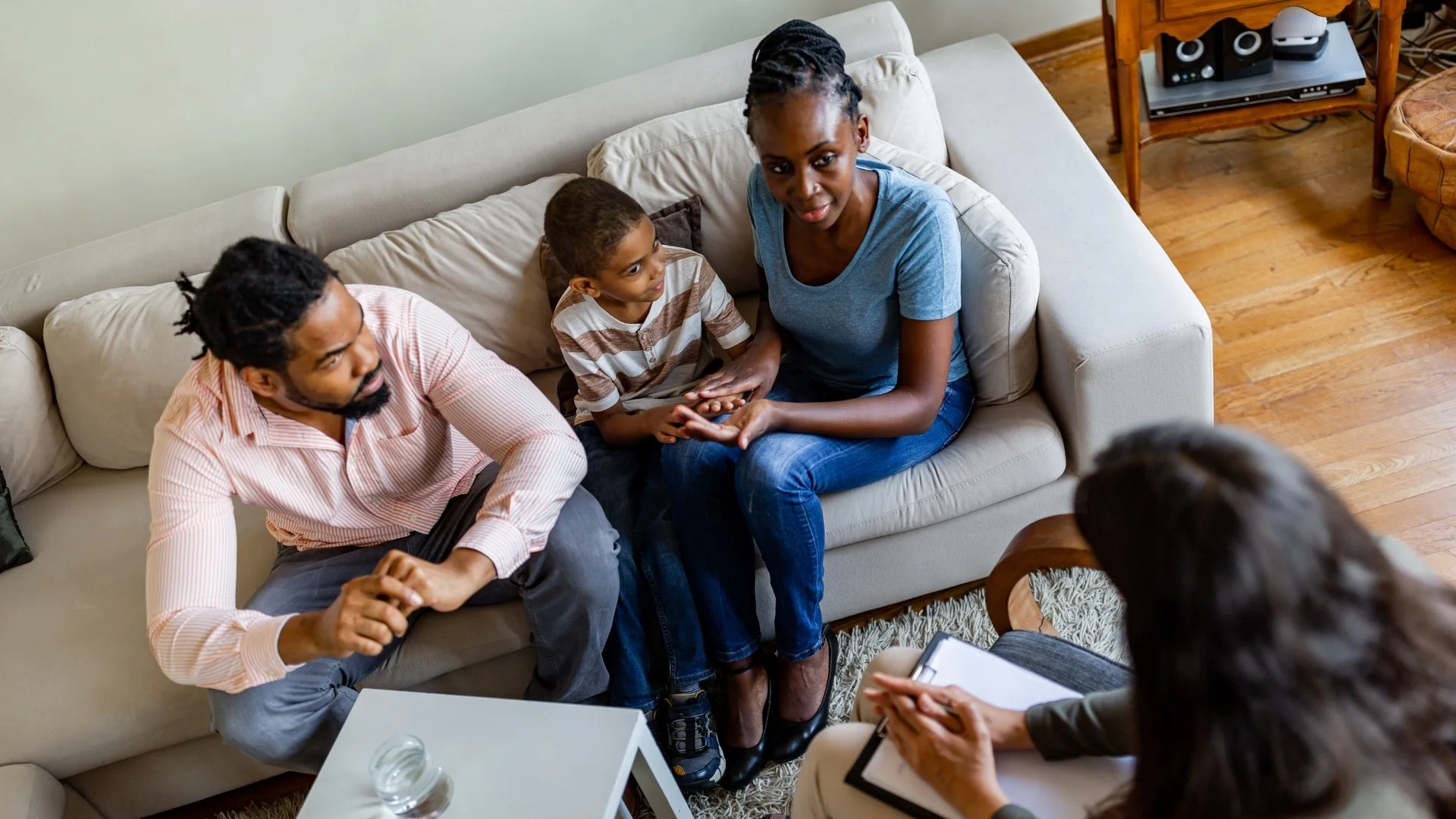 A family of three sits on a couch, talking with a counselor or therapist in a living room setting.