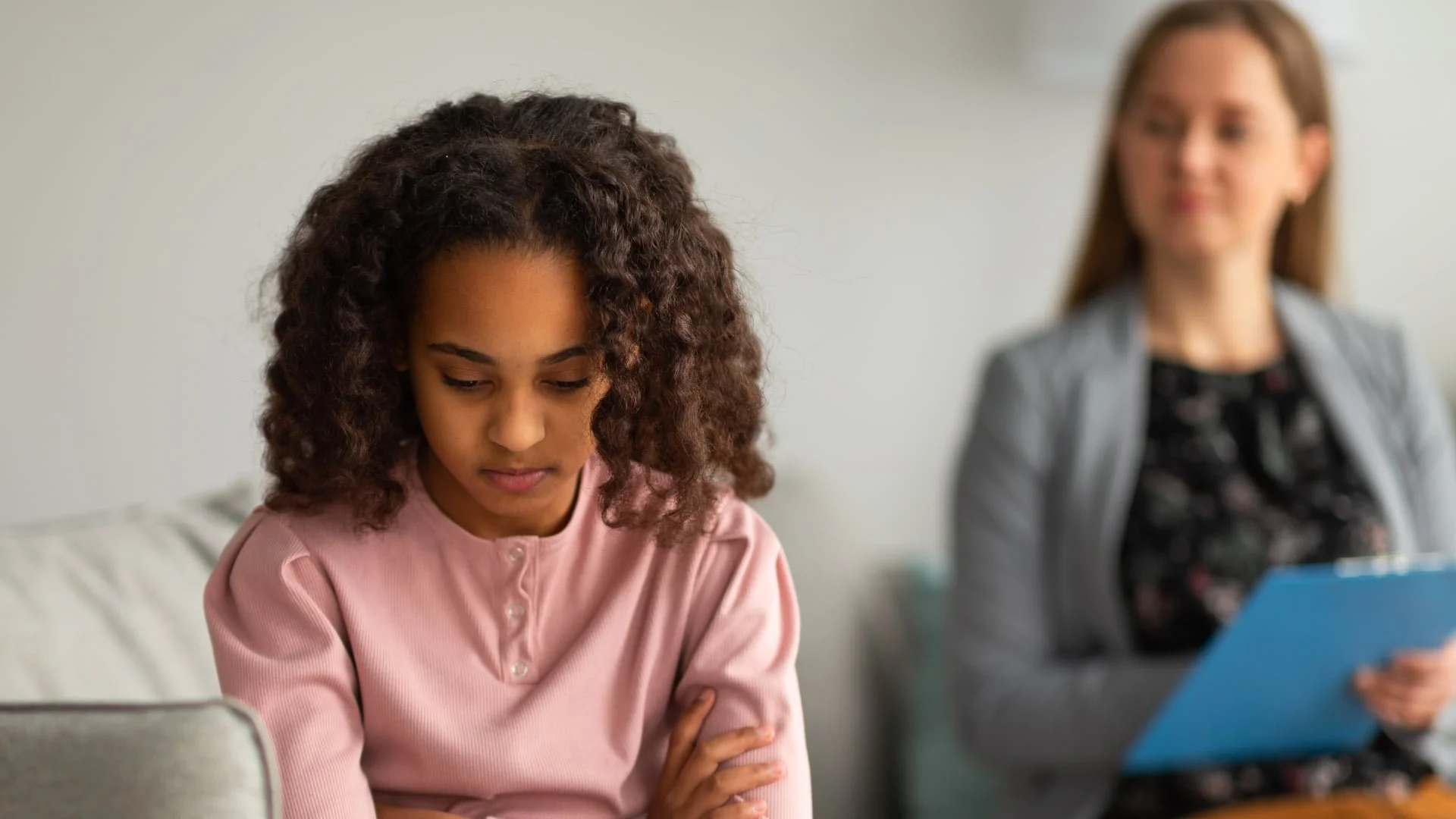 A young girl sits with arms crossed, looking down, while a woman in the background holds a clipboard and observes her.