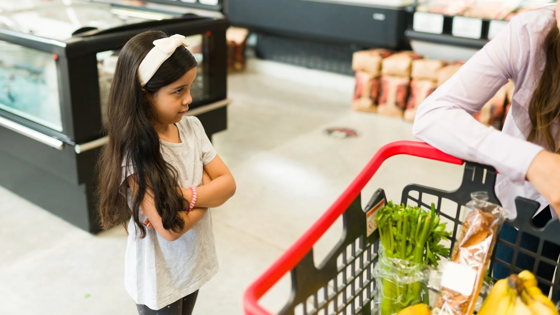 Young girl with long hair and a headband stands with crossed arms, looking at a person shopping in a grocery store.