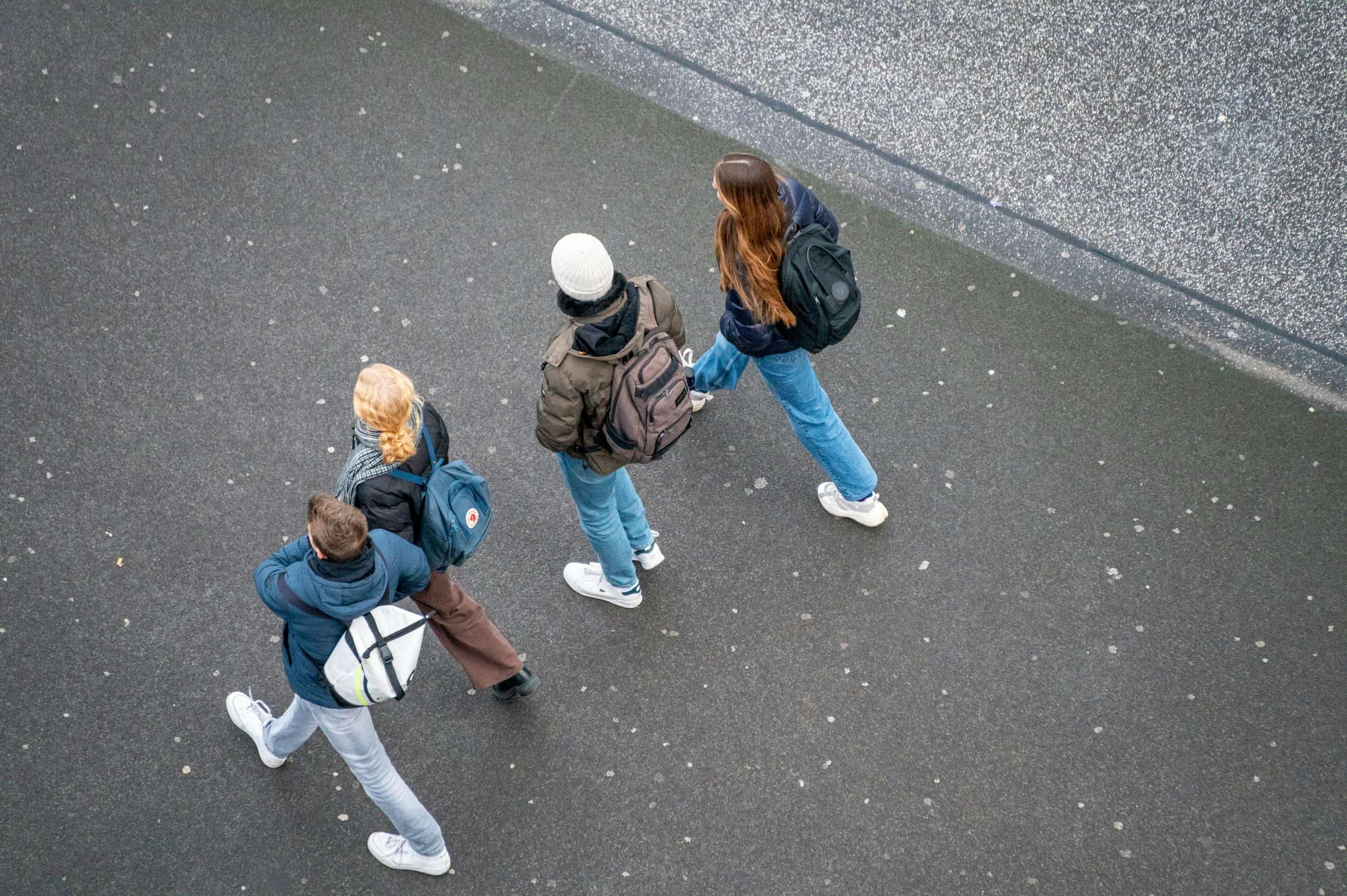 Four students walking on a paved outdoor surface with some snow, carrying backpacks and wearing winter clothing.