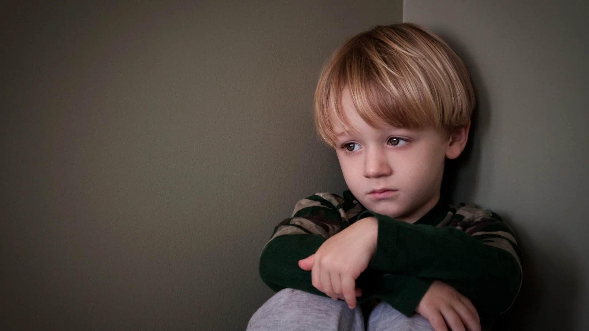 A young boy with blond hair sits alone in a corner, looking sad and thoughtful with his arms wrapped around his knees.