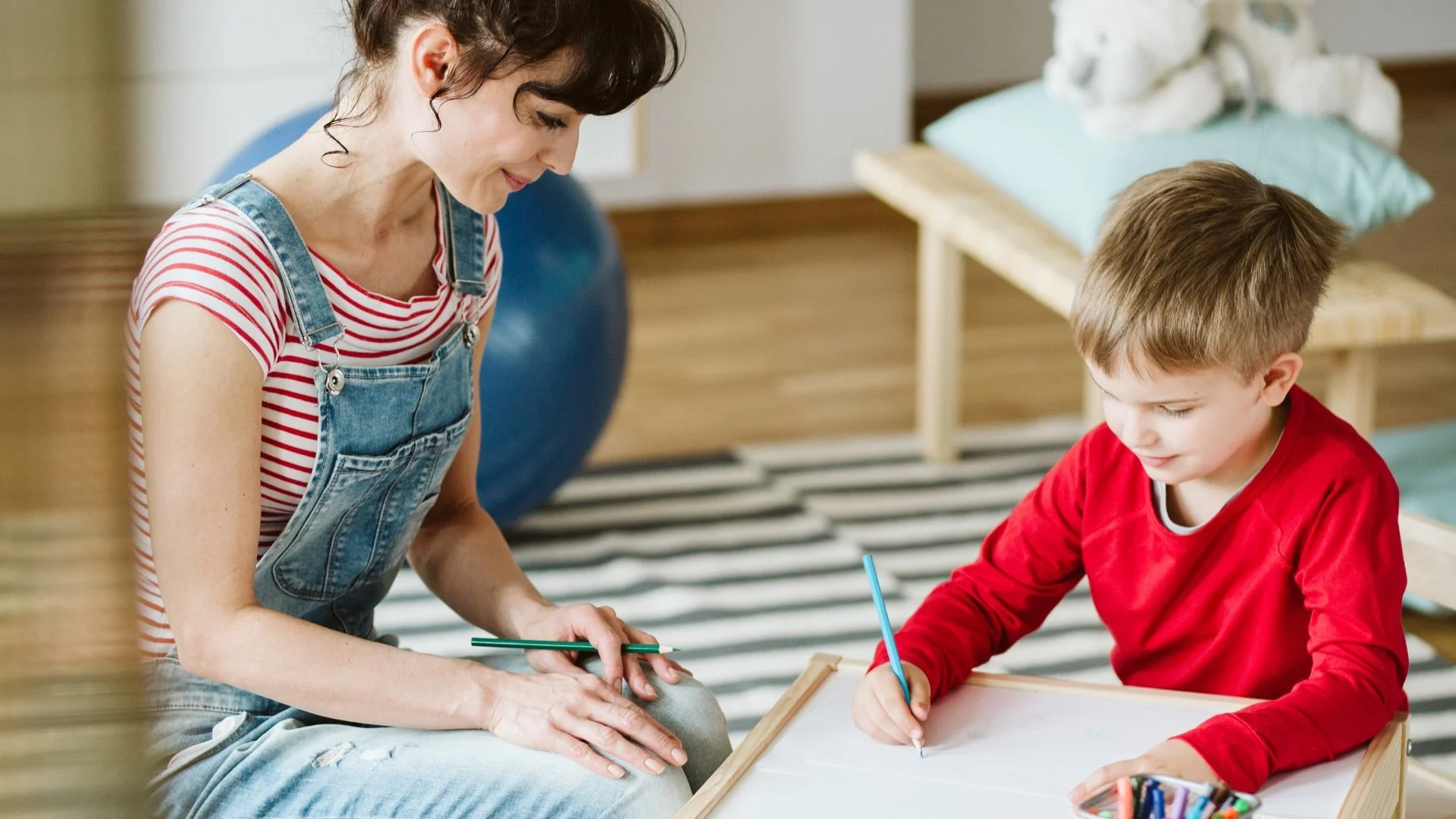 Woman in overalls sits beside a young boy in a red shirt as he draws with colored pencils on a table in a cozy room.