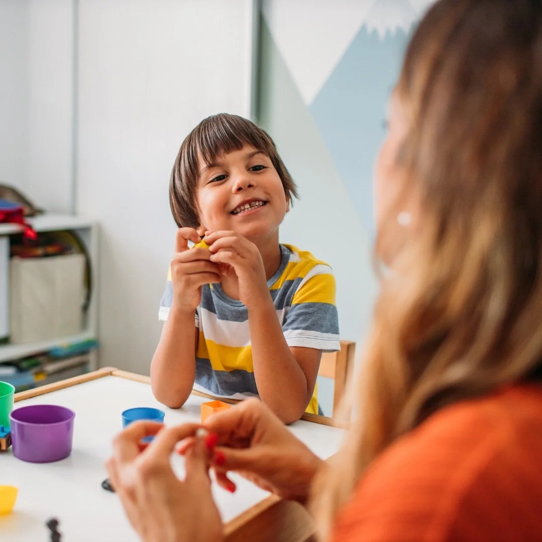Smiling child playing with colorful clay at a table, facing an adult with long hair who is also shaping clay.