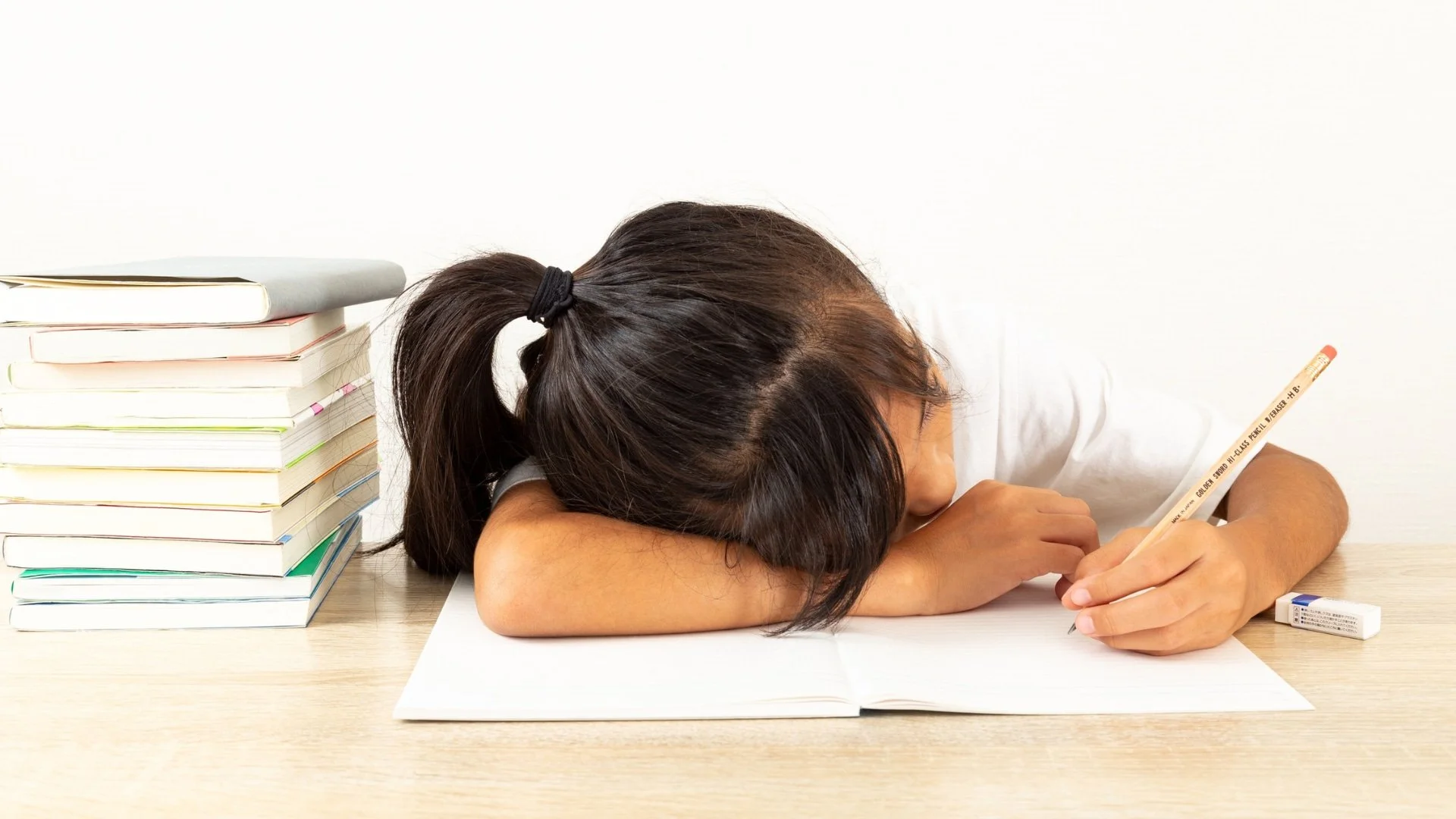A tired student with head down writes in a notebook, sitting beside a stack of books on a desk.