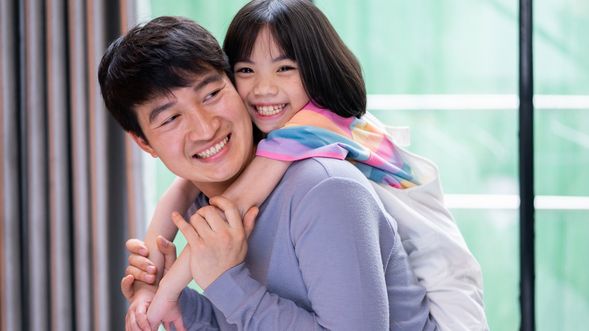 Smiling man gives a piggyback ride to a cheerful young girl indoors, both looking happy and playful.