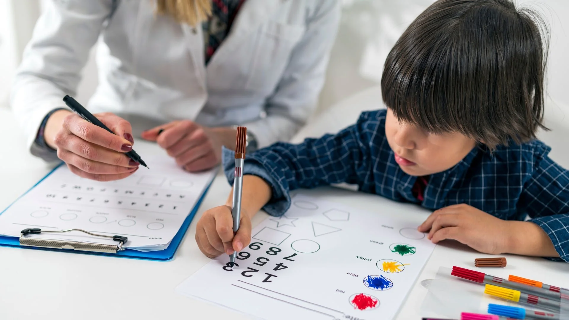 A child completes a worksheet with shapes and numbers beside an adult who is writing on a clipboard.