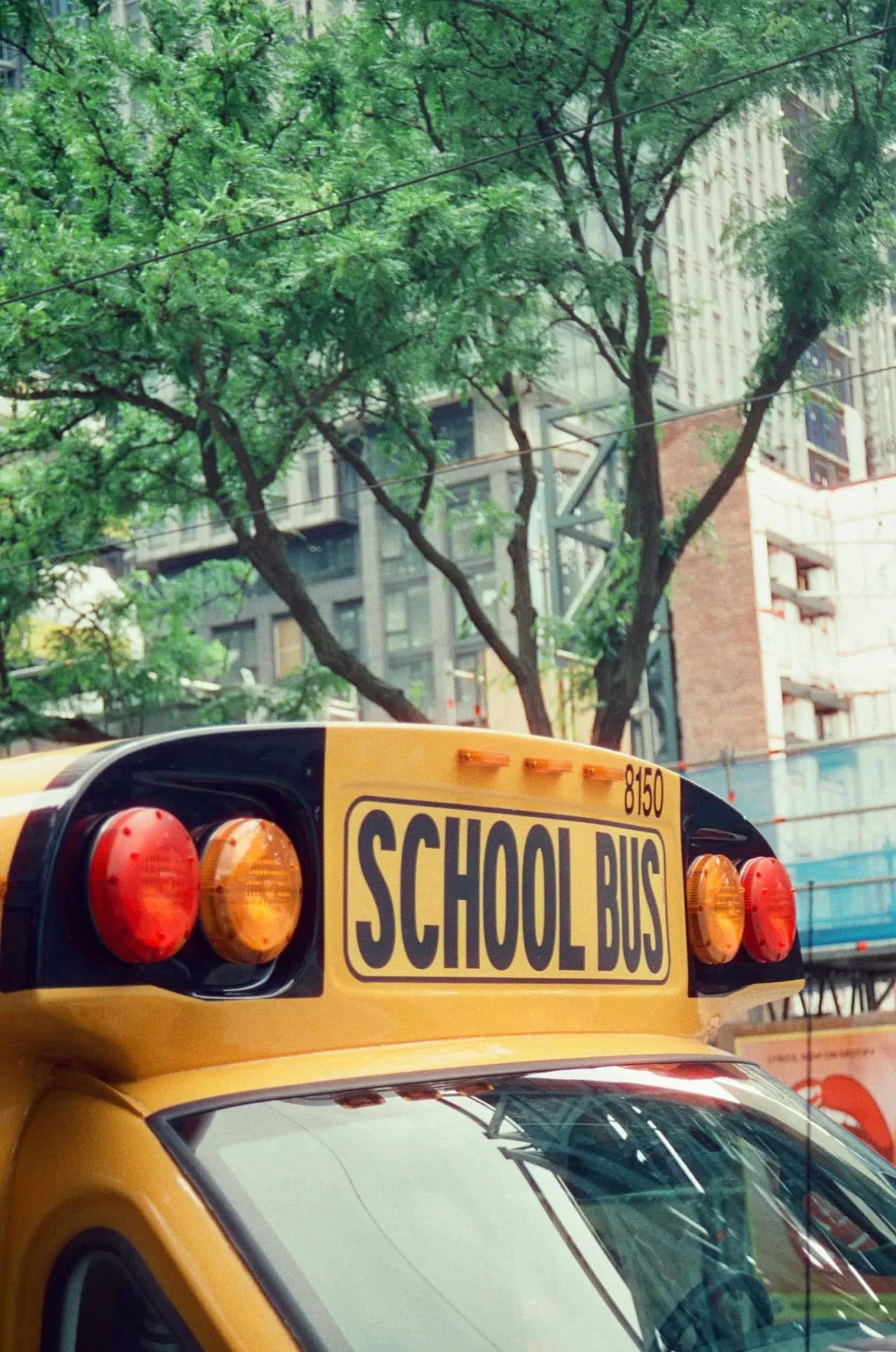 A yellow school bus with flashing lights is parked under green trees in an urban area with tall buildings in the background.