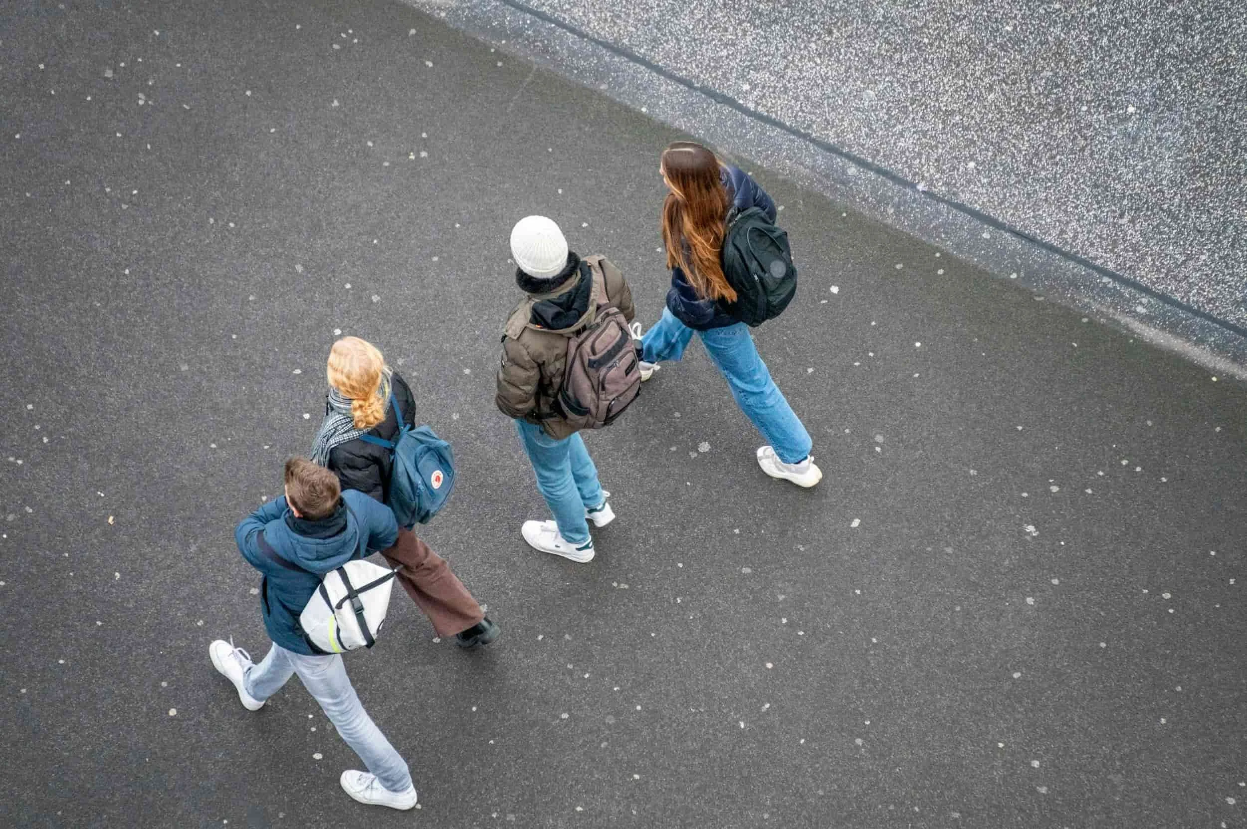 Four people with backpacks walk together on a paved surface, viewed from above.
