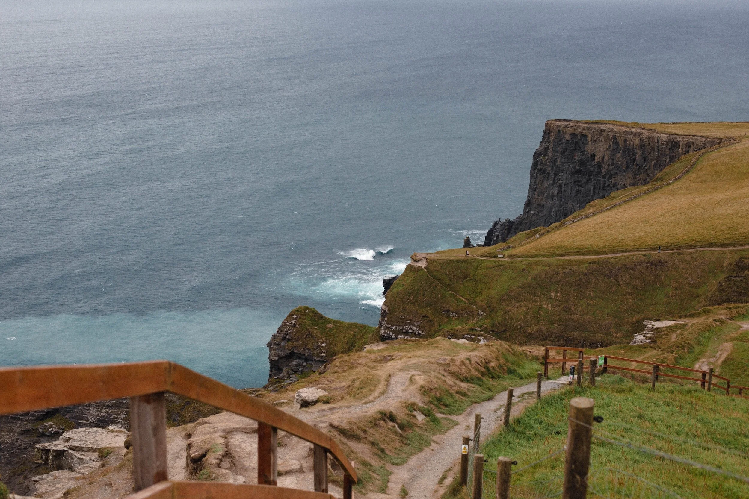 A coastal cliffside trail with wooden railing overlooking the ocean and green grassy slopes.