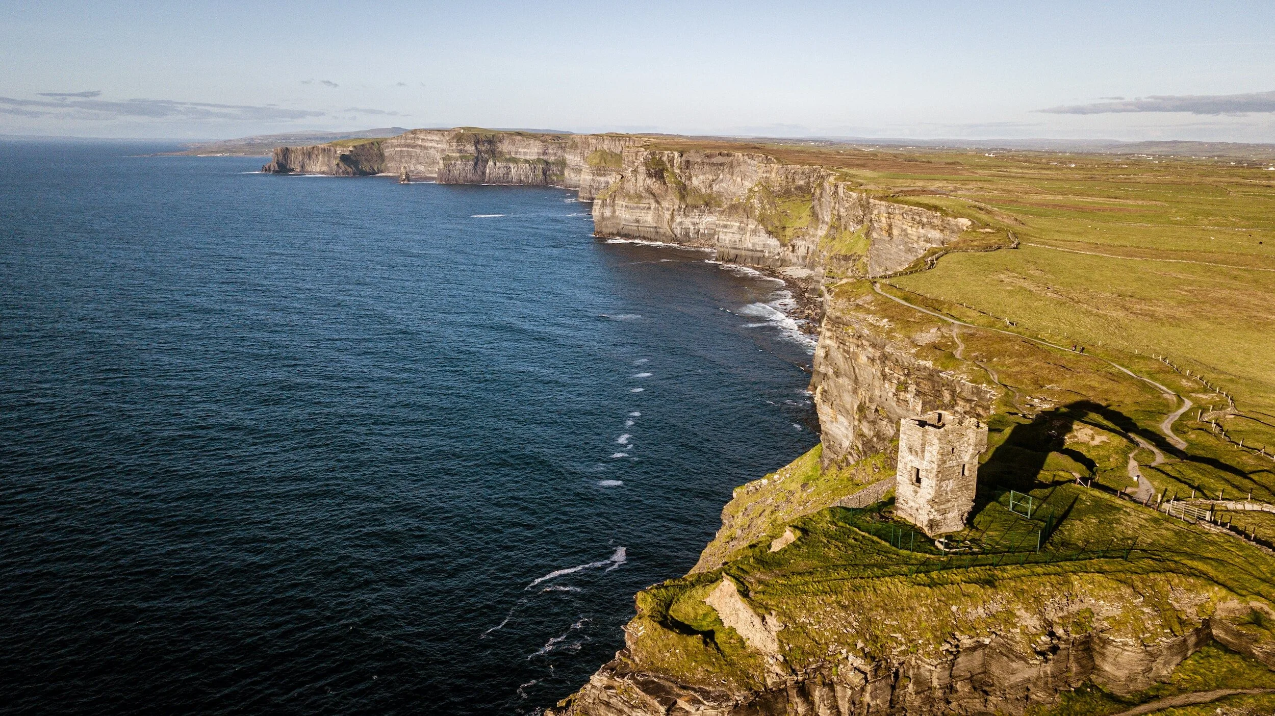 Aerial view of green grassy cliffs along the ocean with a historic stone tower on the right cliff