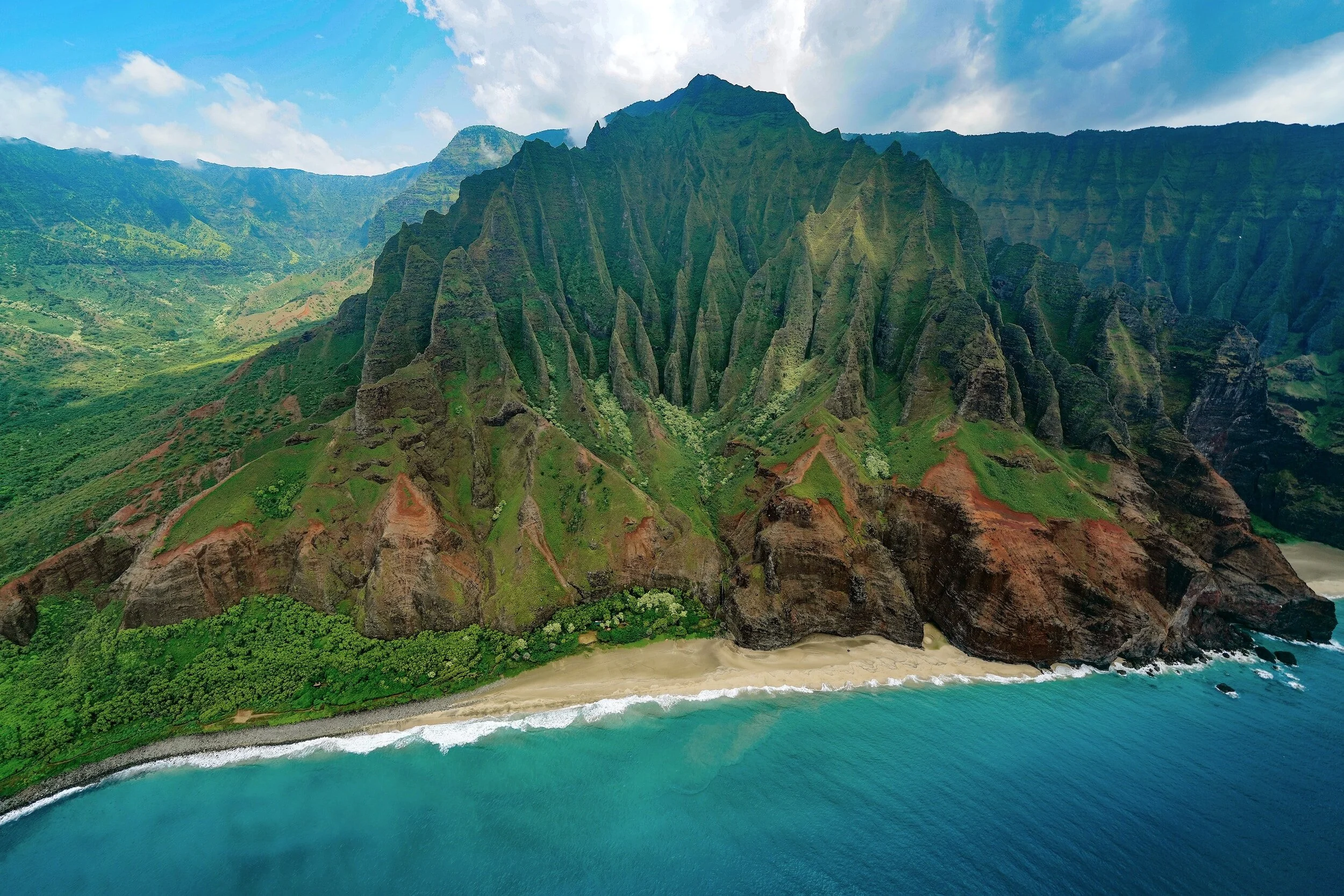 Aerial view of lush green mountains with steep ridges along a sandy beach and turquoise ocean under a partly cloudy sky. Napali Coast, Kauai, Hawaii.