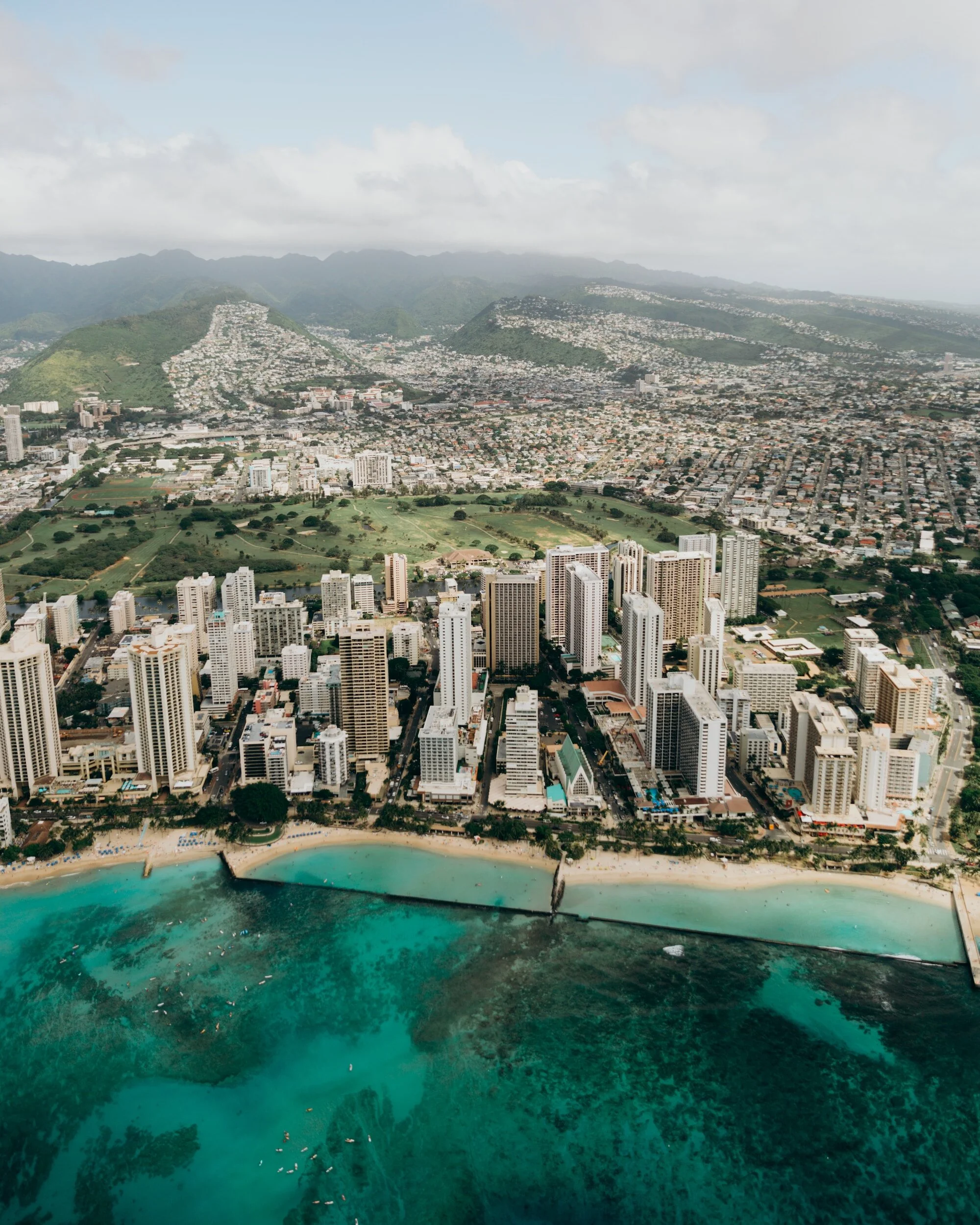 Aerial view of a city with tall buildings along a beach, with green hills and mountains in the background.  Honolulu, Hawaii.