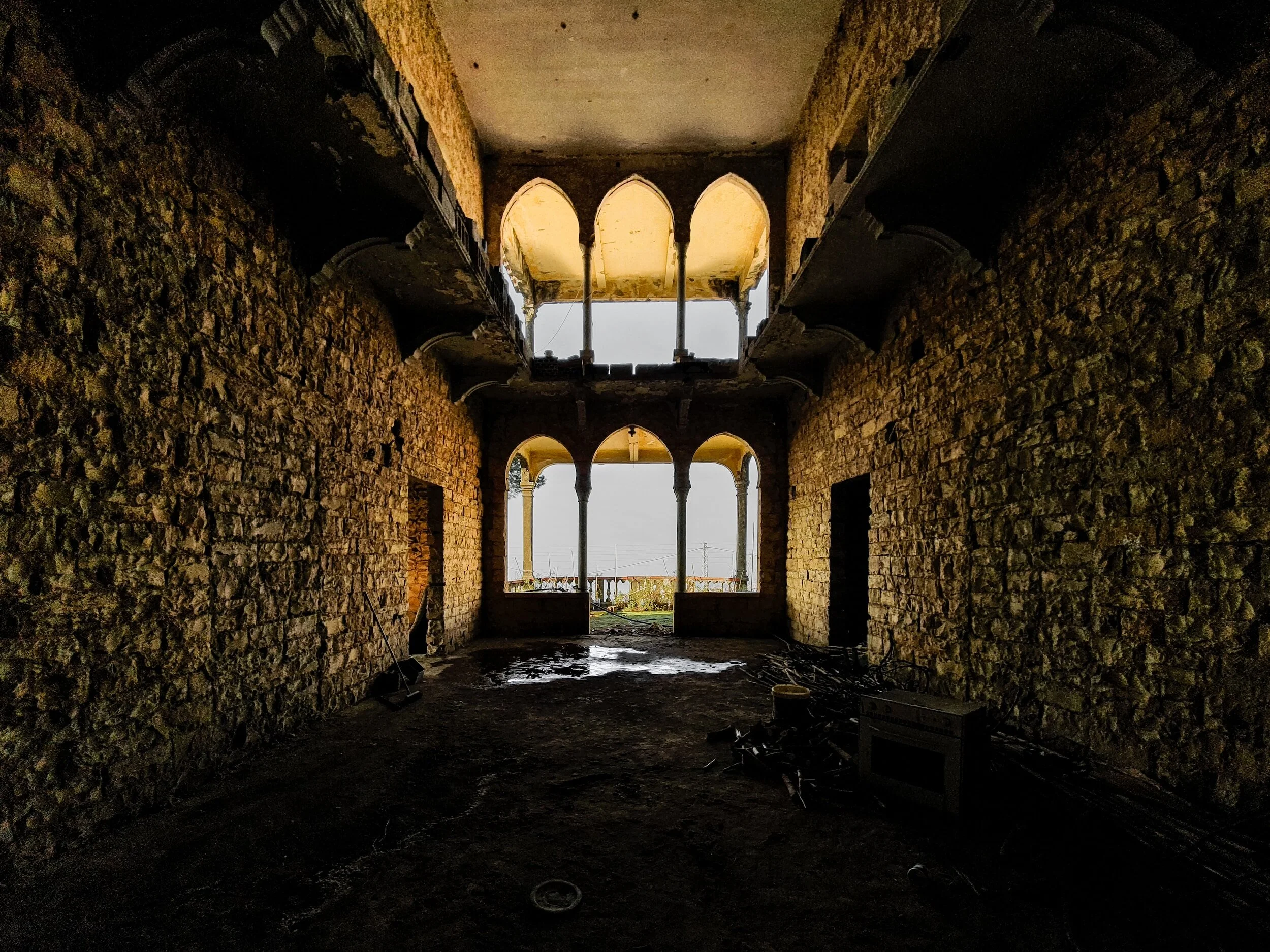 Interior of a stone-building under construction, with arched openings and exposed brick walls, viewed from inside toward open windows showing outdoor scenery.