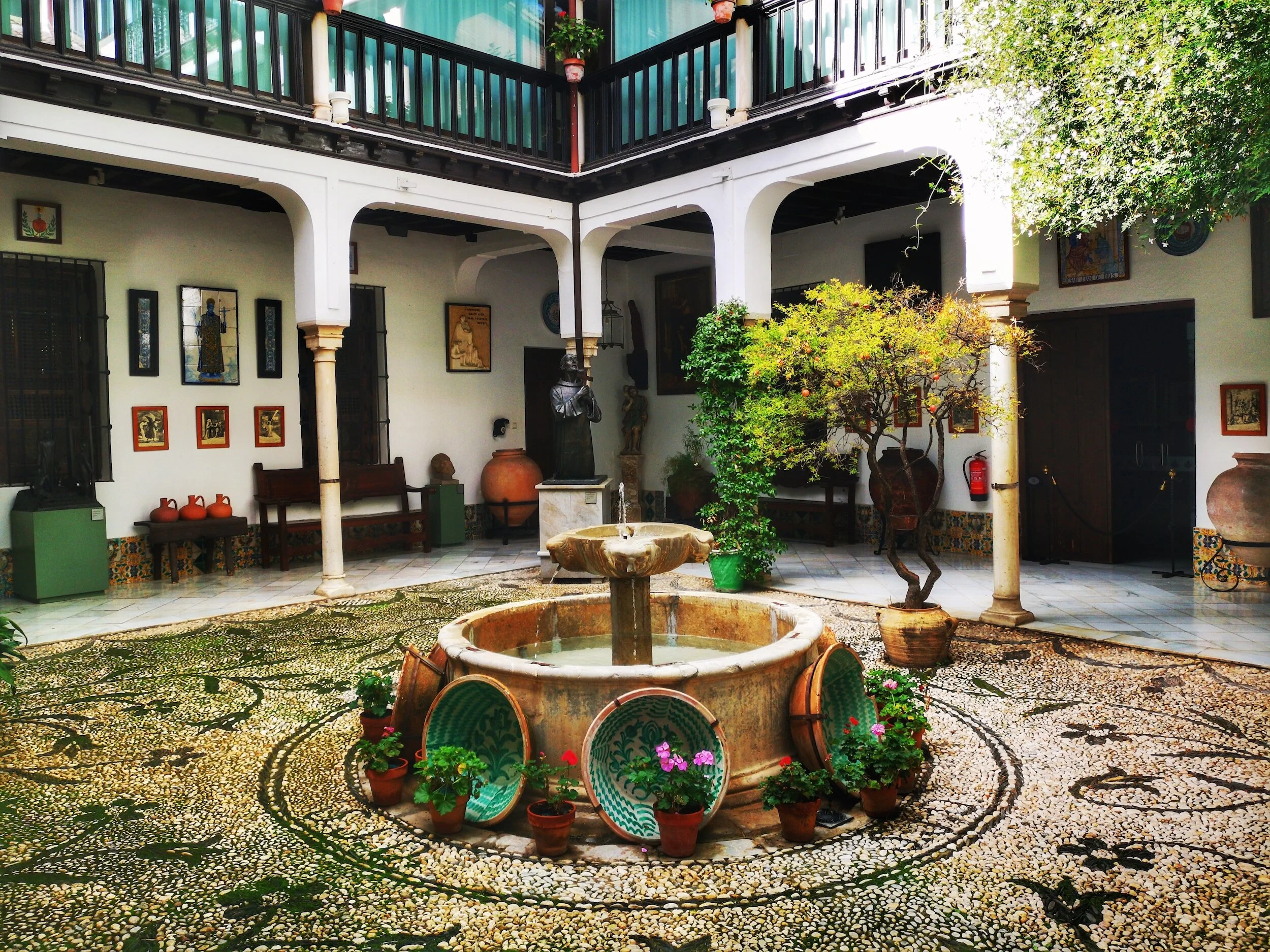 Central courtyard with a fountain surrounded by potted plants, clay pots, and decorative tiles, enclosed by a two-story building with white walls, arches, and a balcony with green curtains and flower pots.
