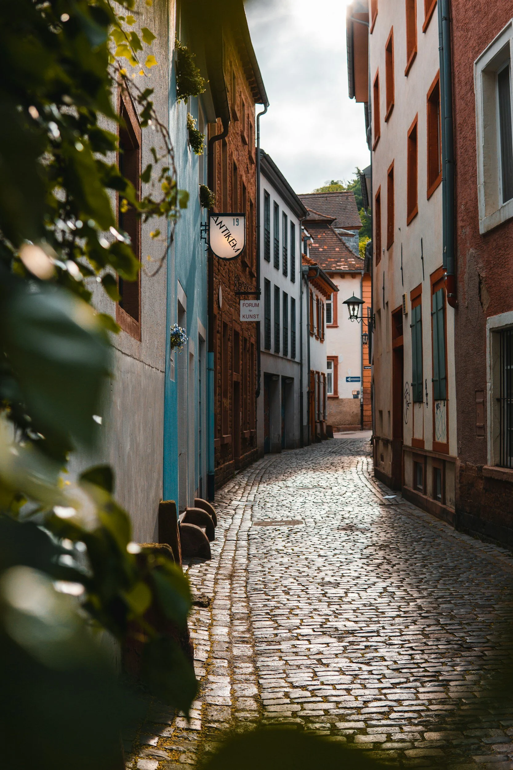 A narrow cobblestone street lined with colorful old buildings with shuttered windows and signs, in what appears to be a European town during daylight.