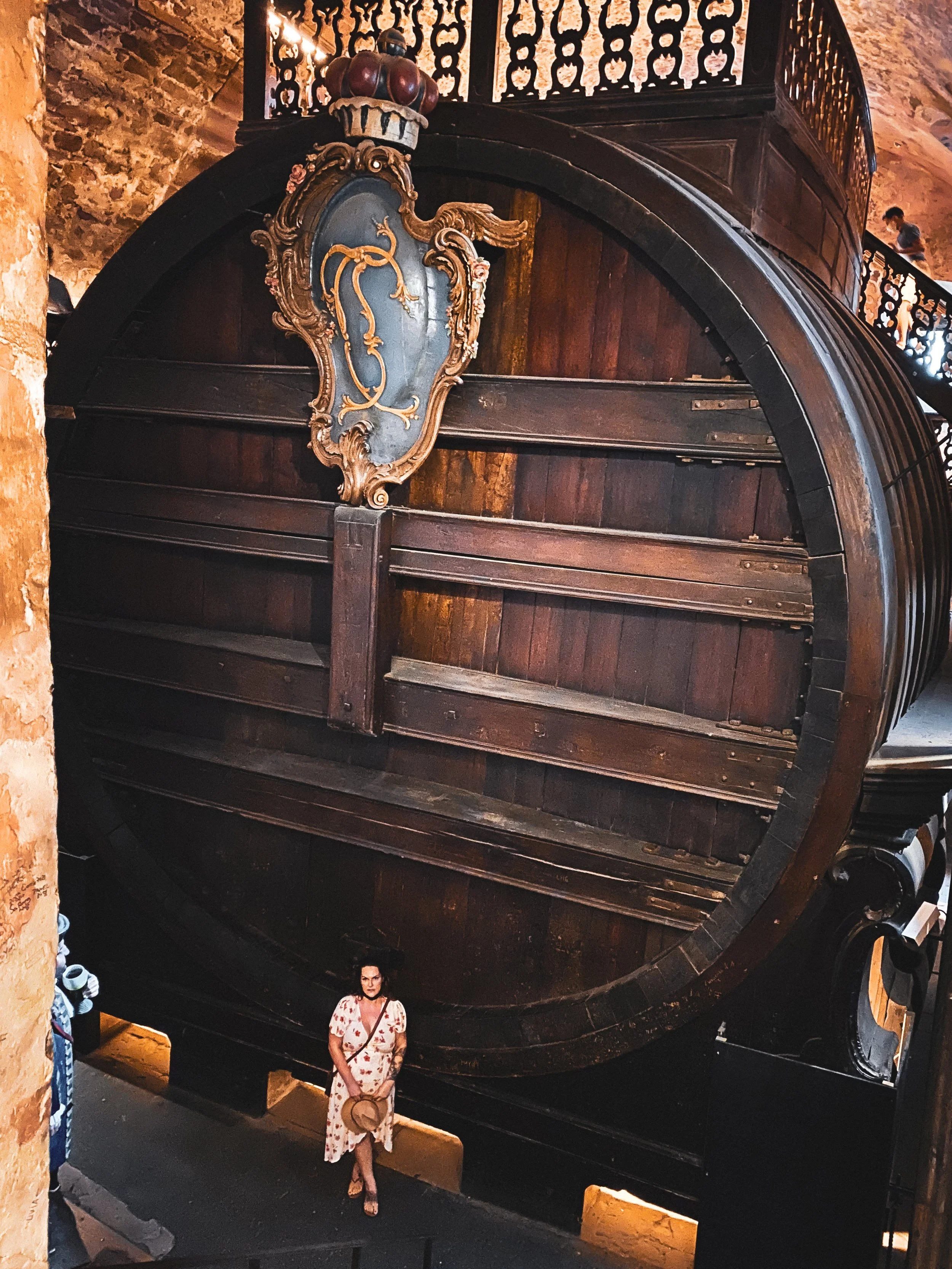 A woman in a floral dress and glasses stands in front of a giant wine barrel in a rustic setting, with stone walls visible in the background.
