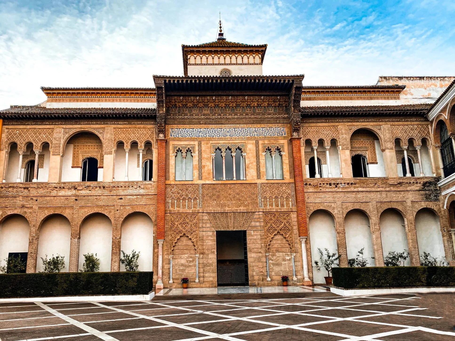 A historic building with intricate architectural details, arches, and decorative elements, featuring a central entrance and a tower-like structure on top, set against a partly cloudy sky.