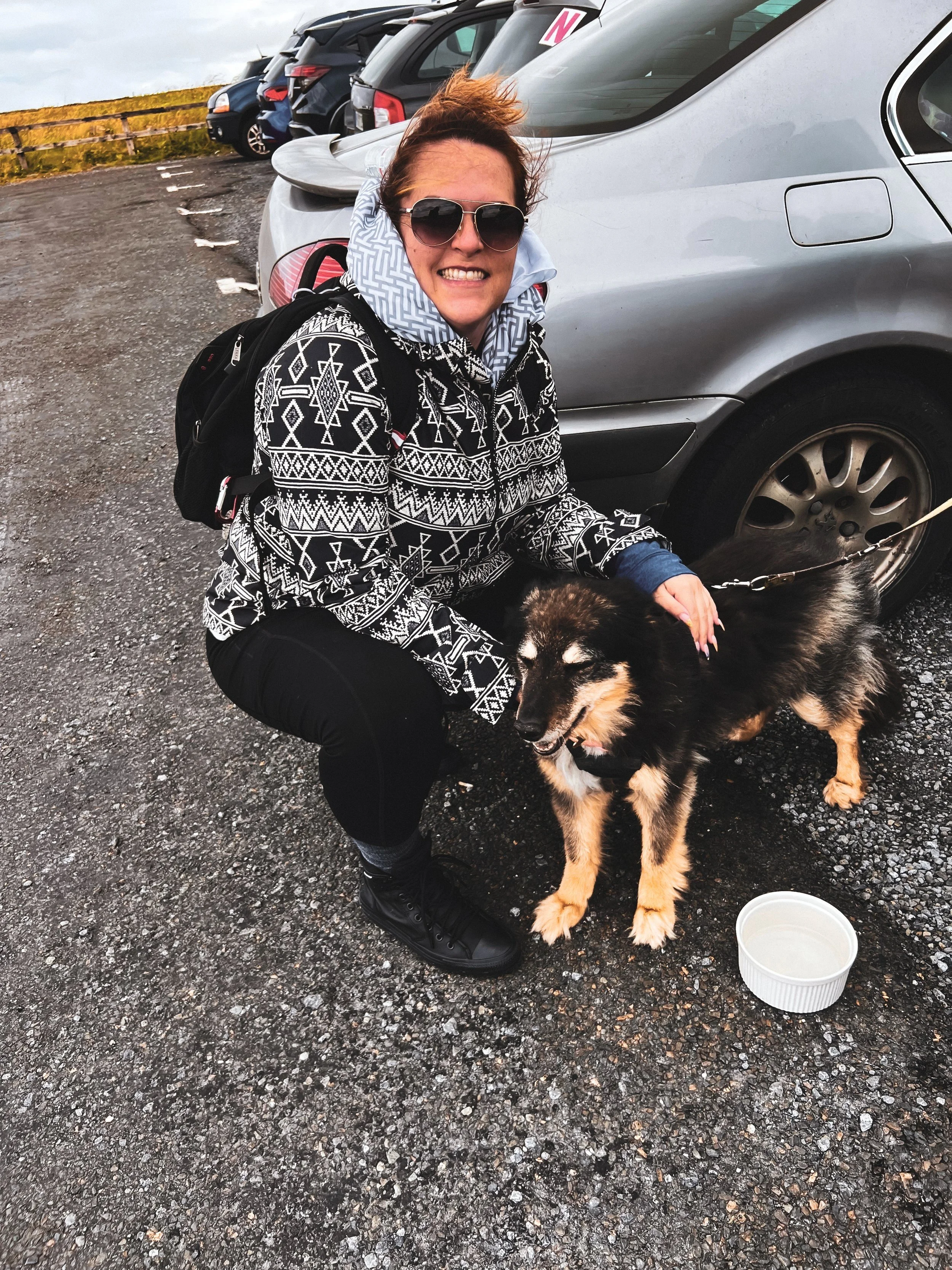 A woman crouching in a parking lot smiling with sunglasses, wearing a black and white patterned jacket, black pants, and black shoes. She has a small black and tan dog on a leash, and there is an empty white bowls on the ground beside them. Multiple parked cars are behind them.