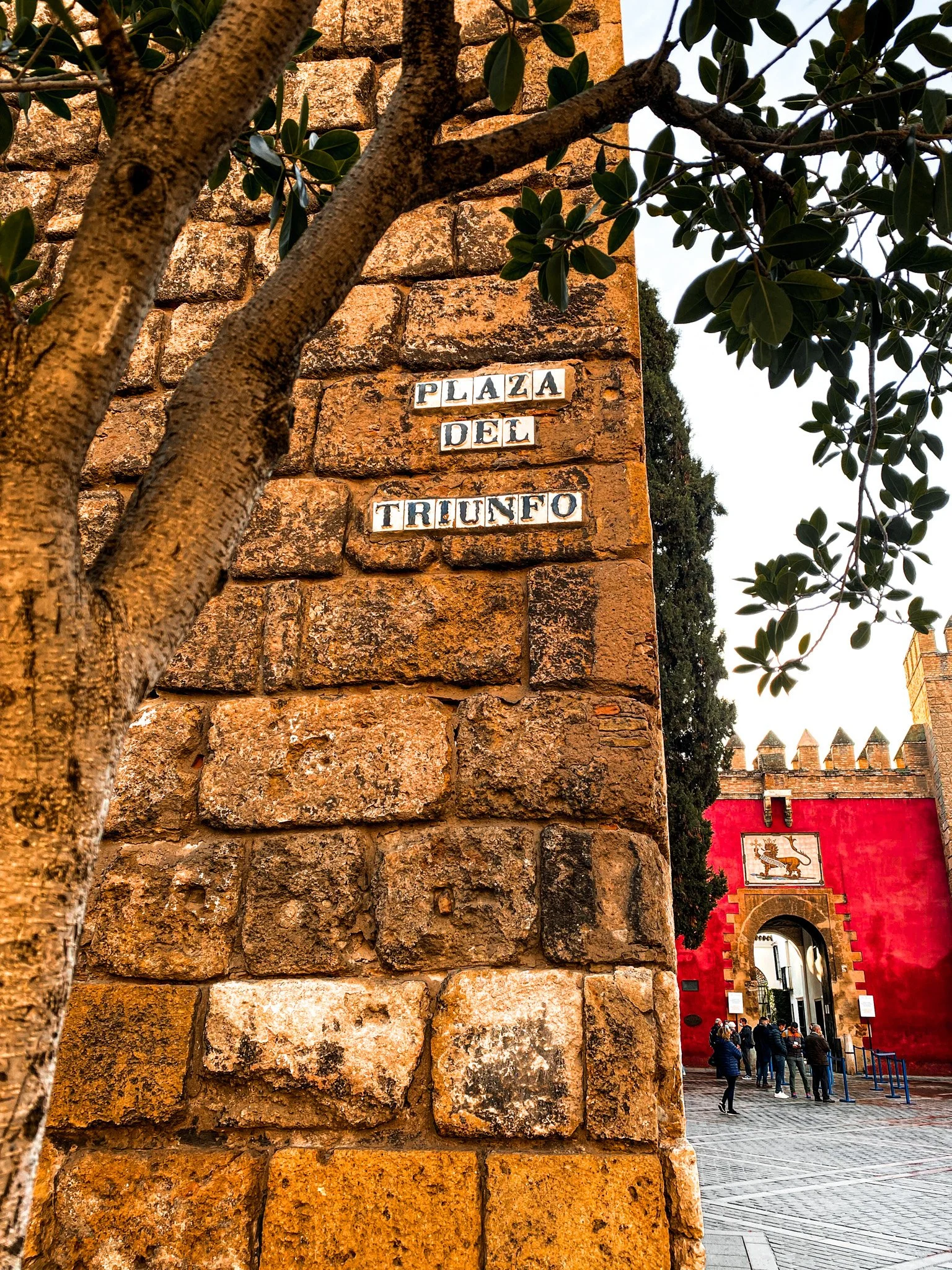 A brick wall with a sign reading 'Plaza del Triunfo,' a red fortification with an arched entrance and a lion crest, and a group of people walking near the entrance.