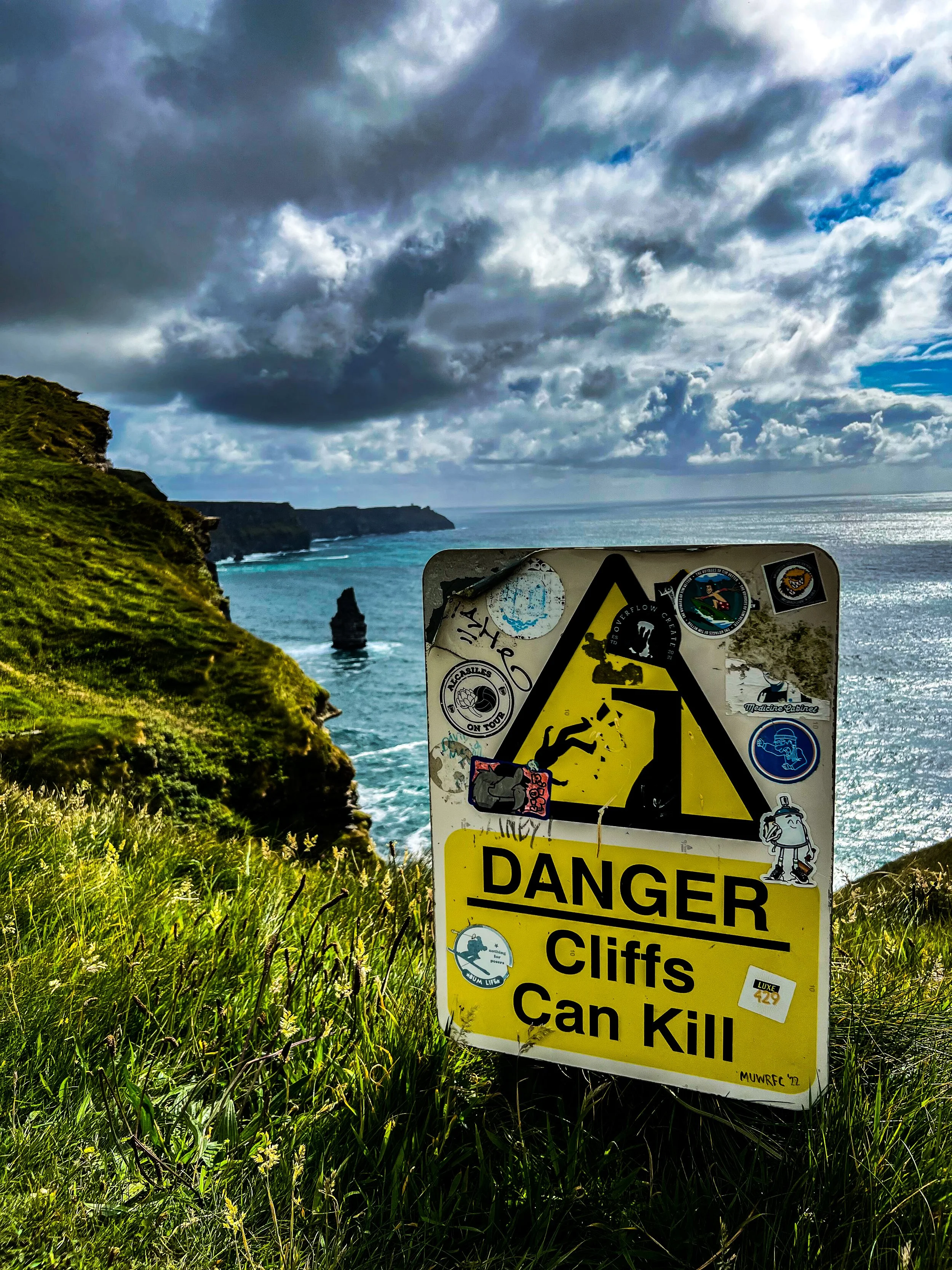 A warning sign on a grassy cliff says 'DANGER Cliffs Can Kill' and has a graphic of a person falling off a cliff. In the background, steep grassy cliffs lead to a rocky shoreline and the ocean under a cloudy sky.