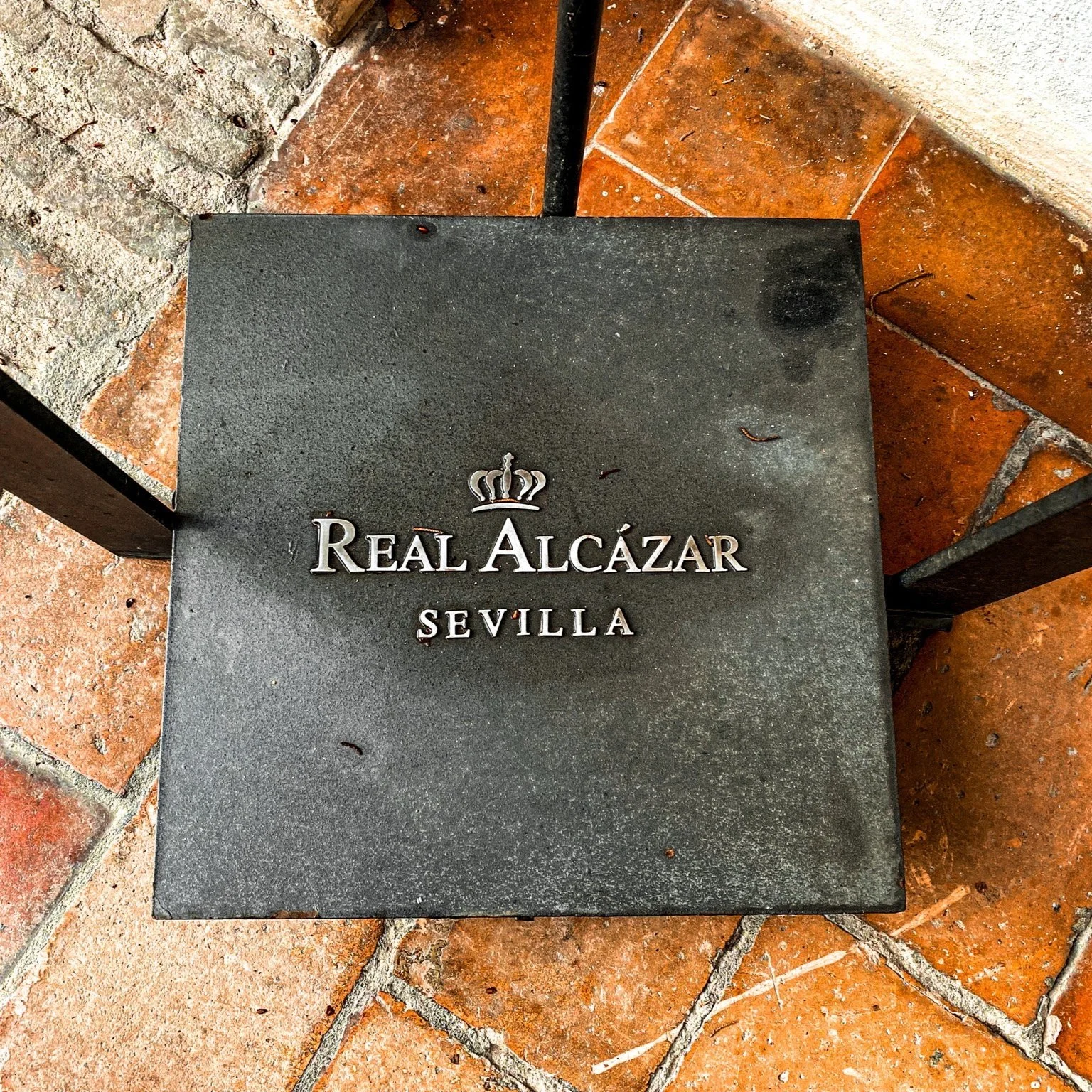 Black sign with silver text reading 'Real Alcázar Sevilla' and a crown symbol, on a tiled floor.