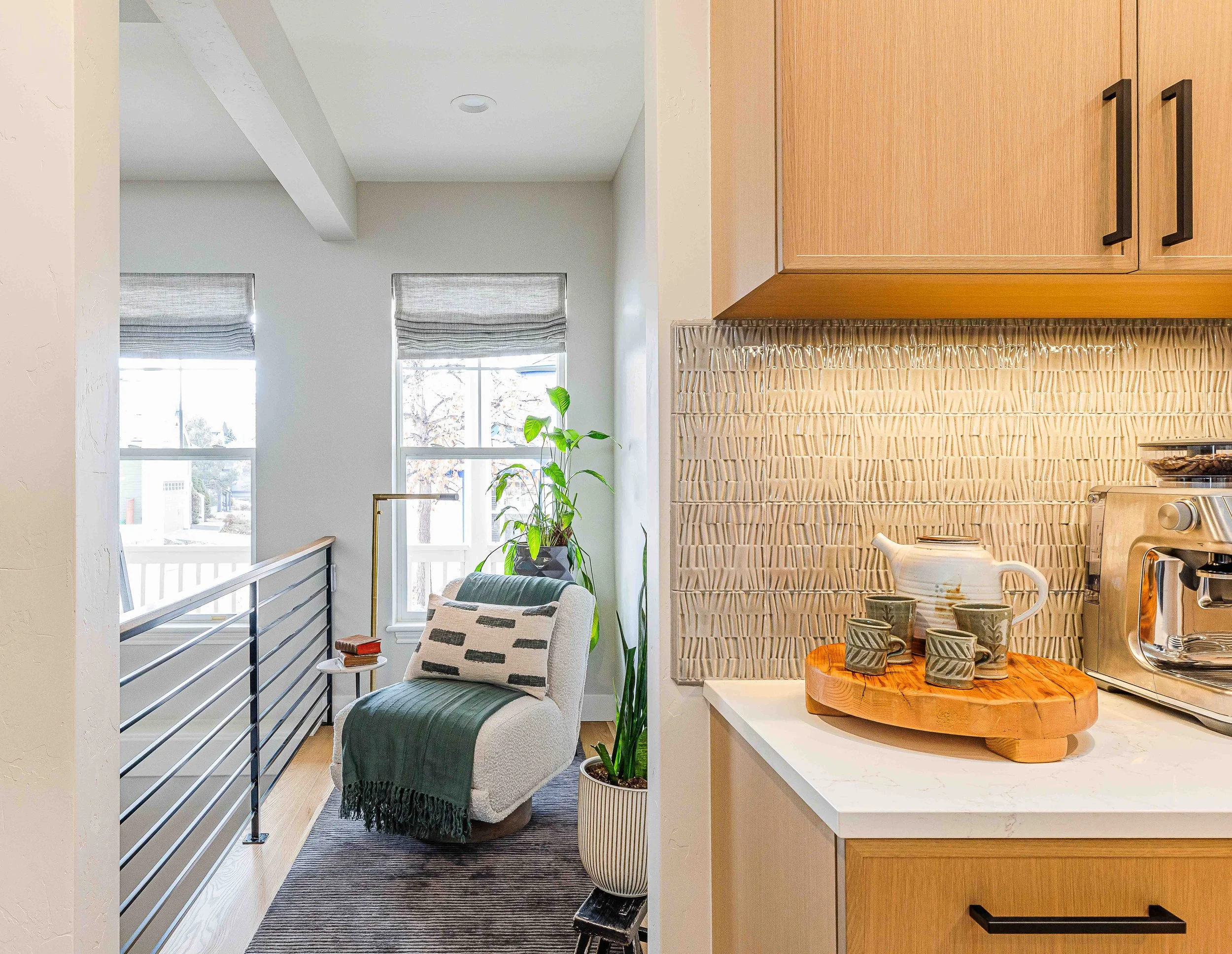 Interior view of a cozy living space adjacent to a kitchen, featuring a white armchair with a green throw and striped pillow, a tall potted plant, windows with gray Roman shades, a small side table, and kitchen cabinets with a textured backsplash dis