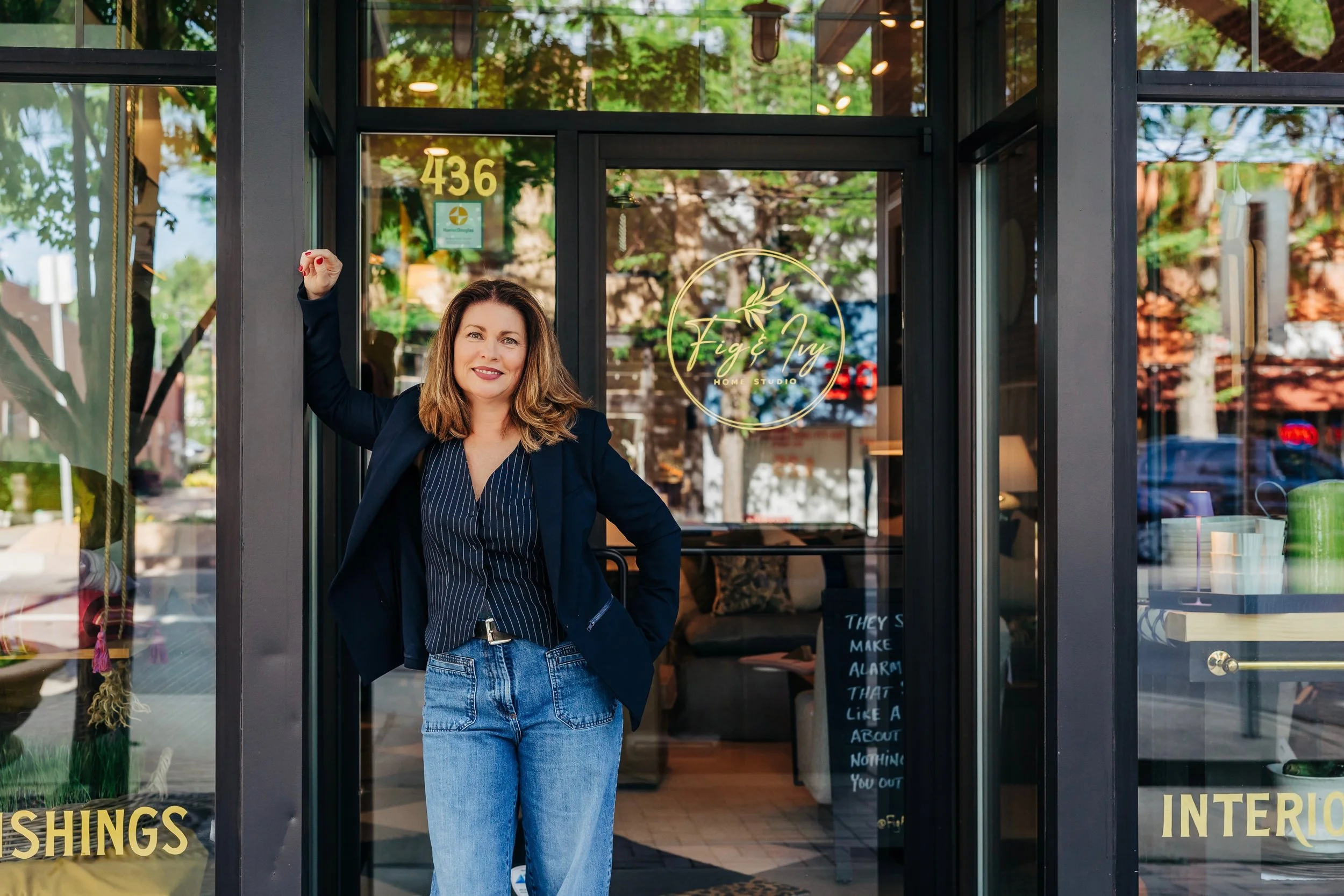 A woman standing in front of a glass storefront door, smiling, with her left arm raised and leaning against the frame. The door has a sign that says 'Fig & Ivy Home Studio' and the numbers 436 above. The interior of the store is visible through the glass, displaying various home decor items.