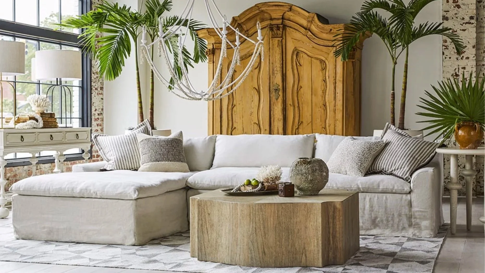 Living room with a white sectional sofa, a large wooden armoire, green tropical plants, a chandelier, a coffee table with decorative items, a white sideboard, and large windows with brick accents.