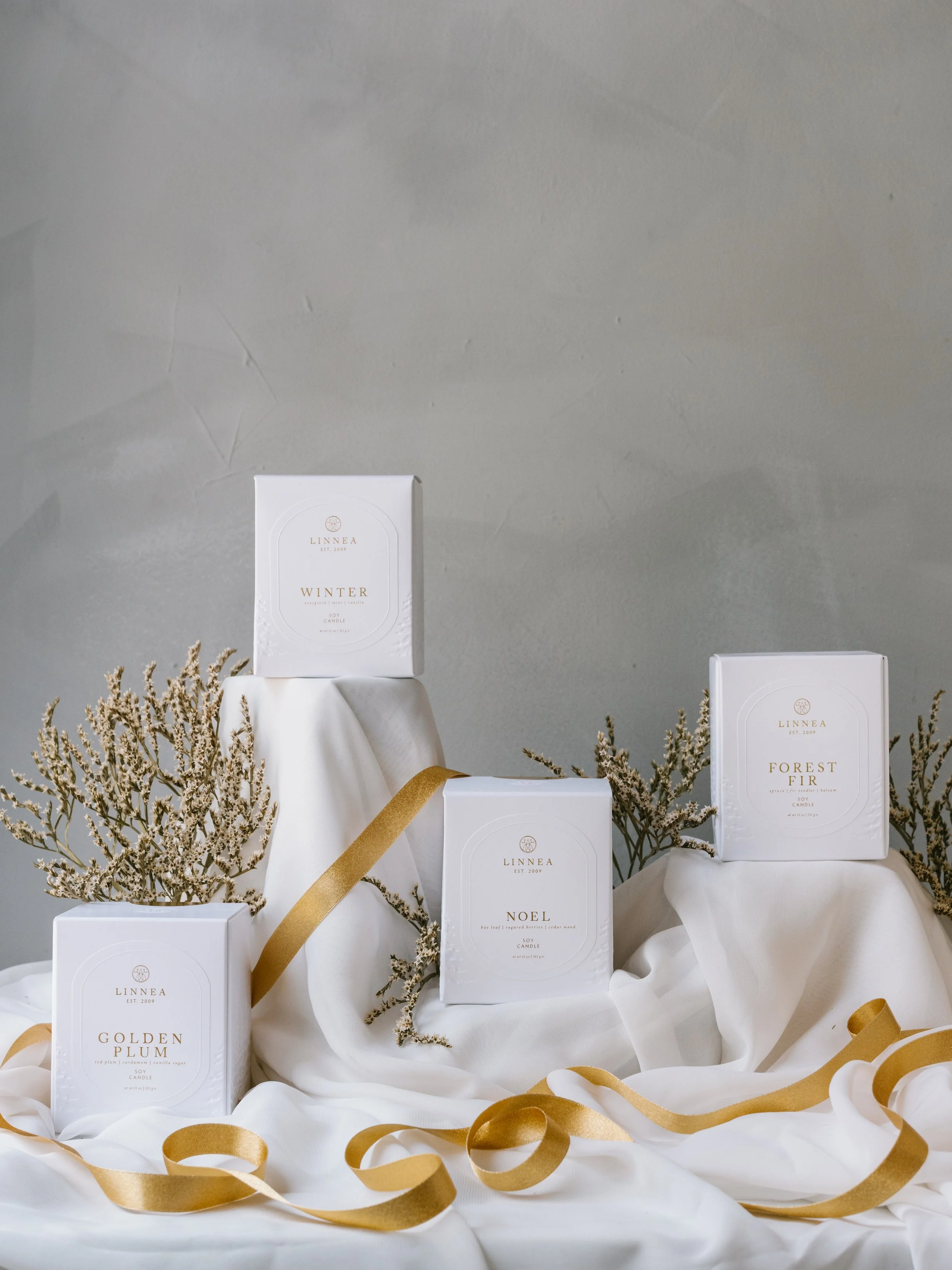 Display of four white boxes of scented candles with beige/white dried flowers and gold ribbons, arranged on white fabric against a gray wall.