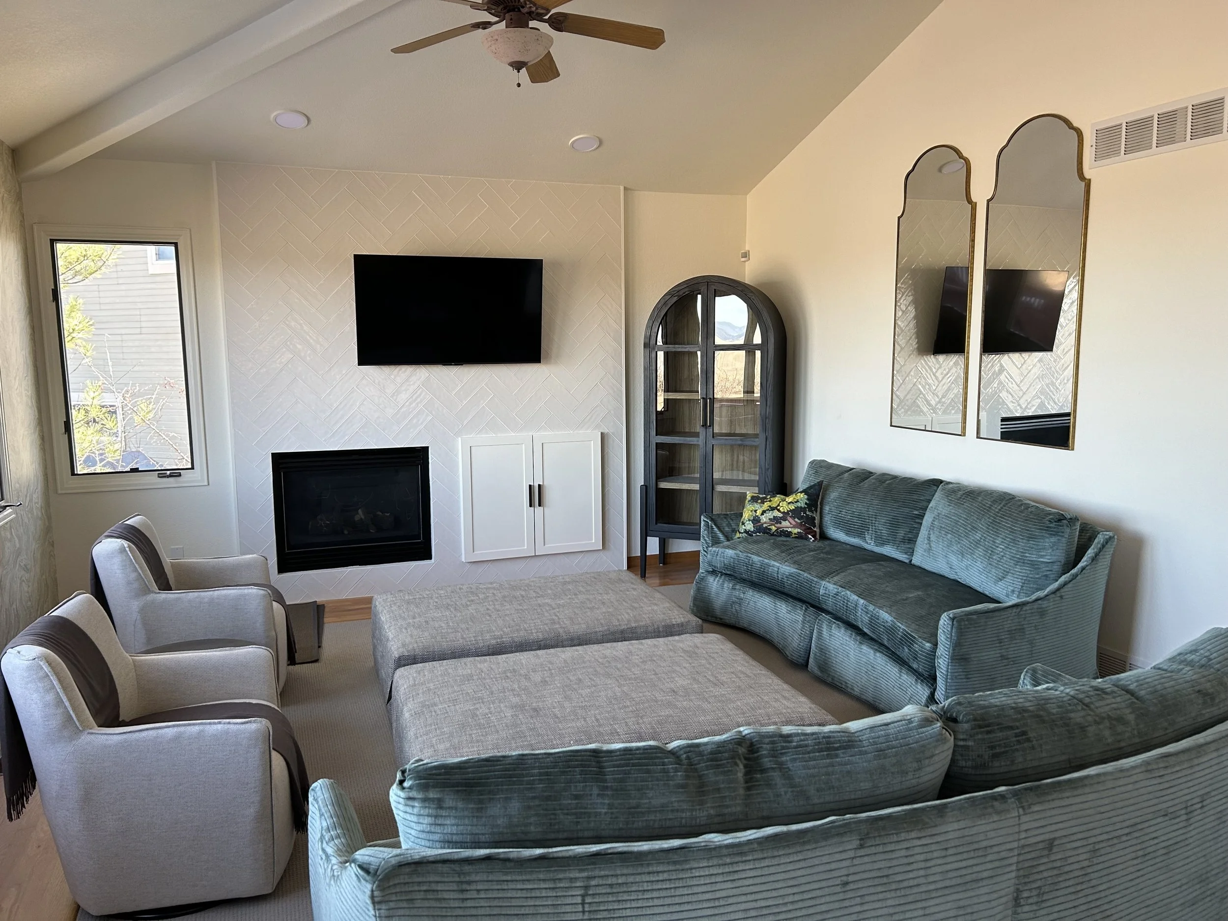 Living room with a teal velvet sofa, two beige armchairs, a large ottoman, a wall-mounted TV, a fireplace, and a window with a view outside.