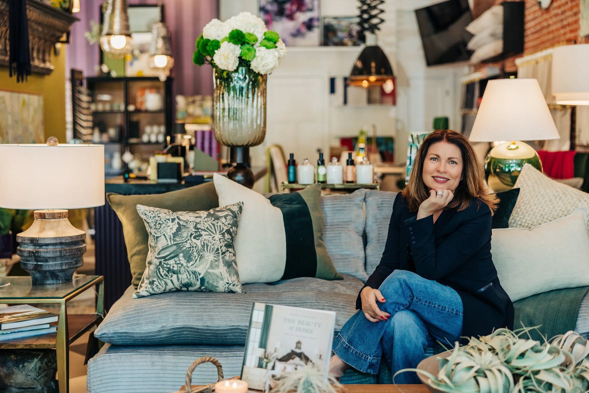 A woman sitting on a sofa in a decorated interior space with lamps, pillows, and a large floral vase, smiling and resting her chin on her hand.