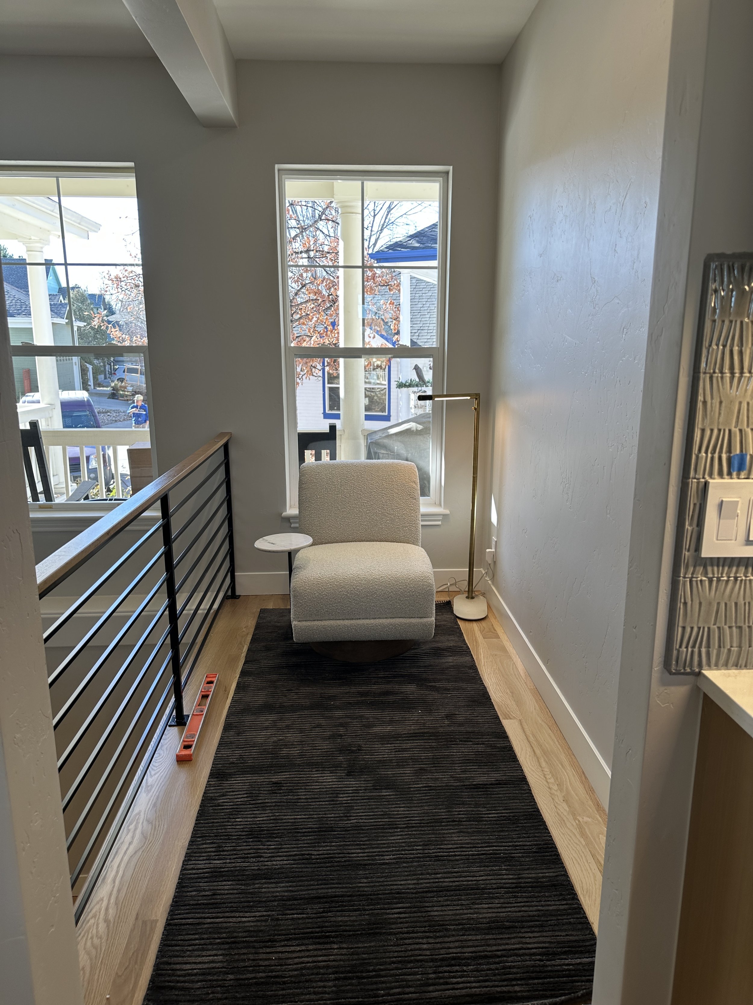 A cozy nook with a modern armchair, a small side table, a floor lamp, and large windows showing trees and neighboring houses outside.