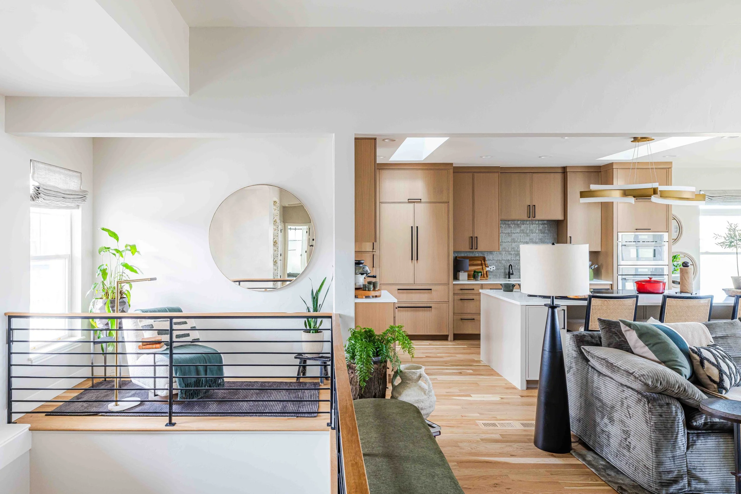 Open-concept living room and kitchen with light wood cabinets, white walls, hardwood flooring, and modern furniture, including a gray sofa, dining chairs, and plants.