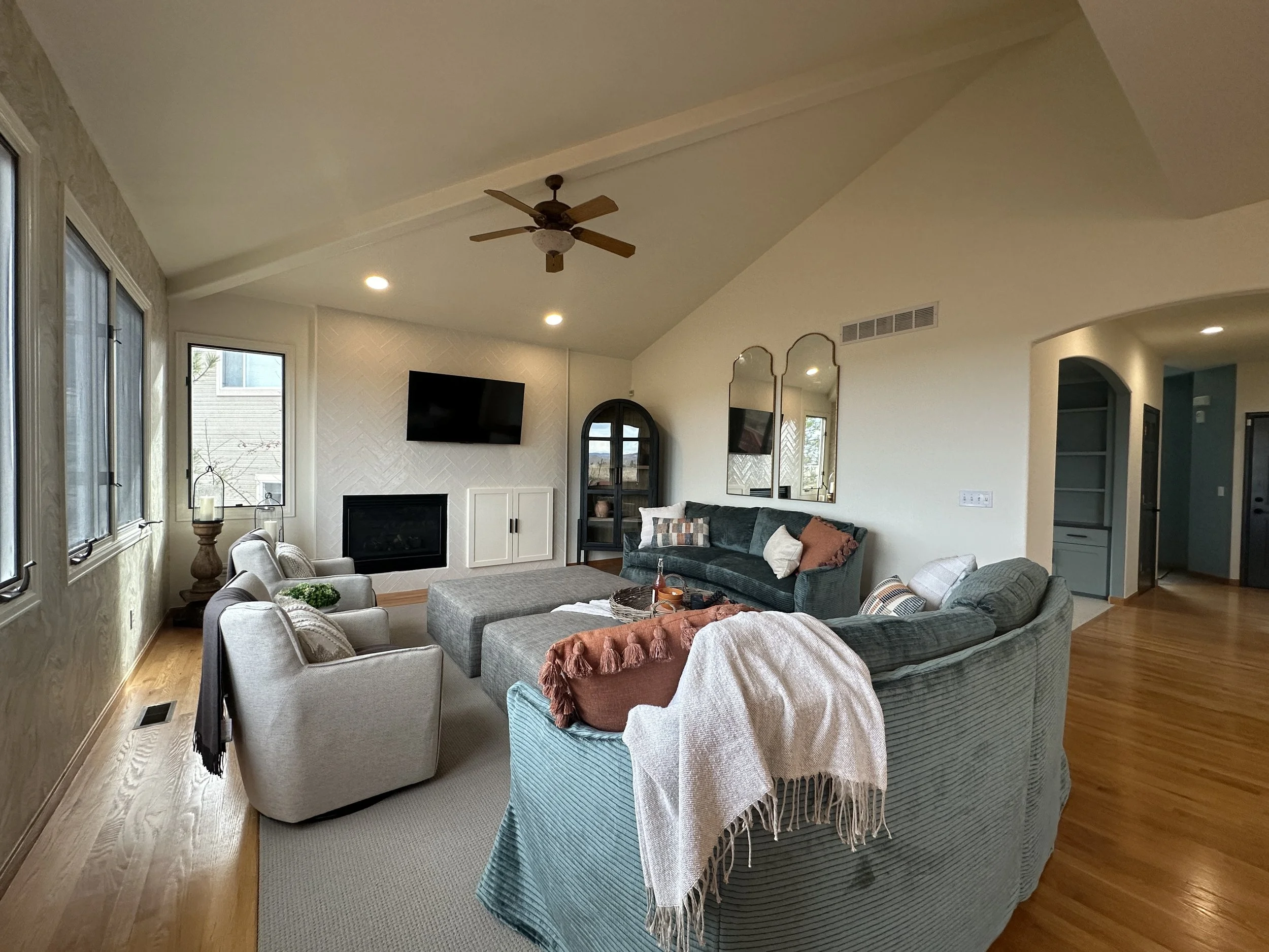 Living room with large sectional sofa, armchair, television above fireplace, windows on left, wooden floors, ceiling fan, decorative mirrors, and a corner cabinet.