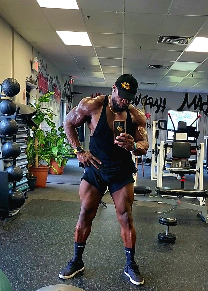 A muscular man in a gym taking a selfie in front of a mirror. He is wearing a black tank top, black shorts, a cap, and Nike socks and shoes. The gym has various exercise equipment and green plants.