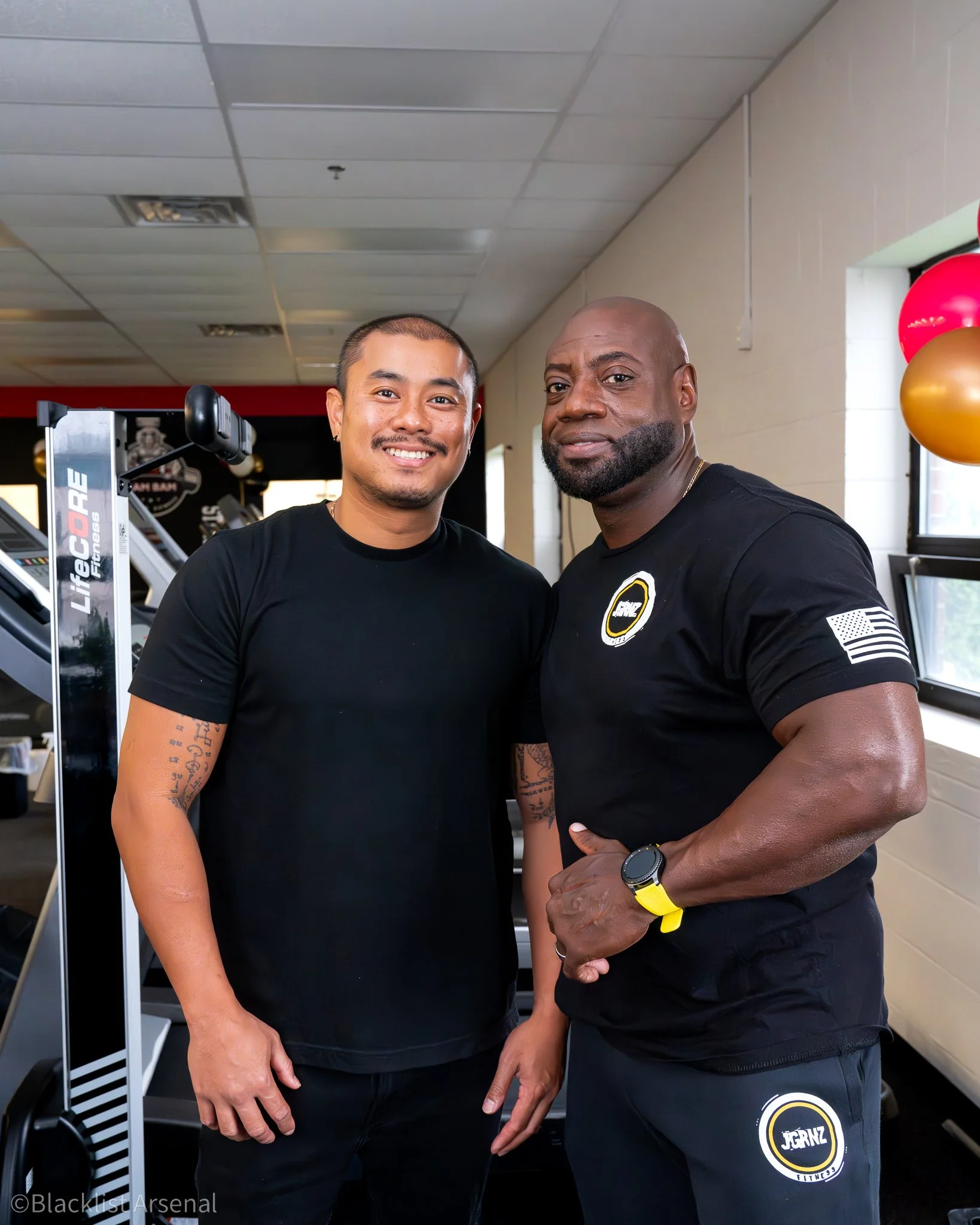Two men standing inside a gym, smiling. One has short dark hair and the other is bald with a beard. They are wearing black shirts with gym logos and are surrounded by gym equipment and balloons.