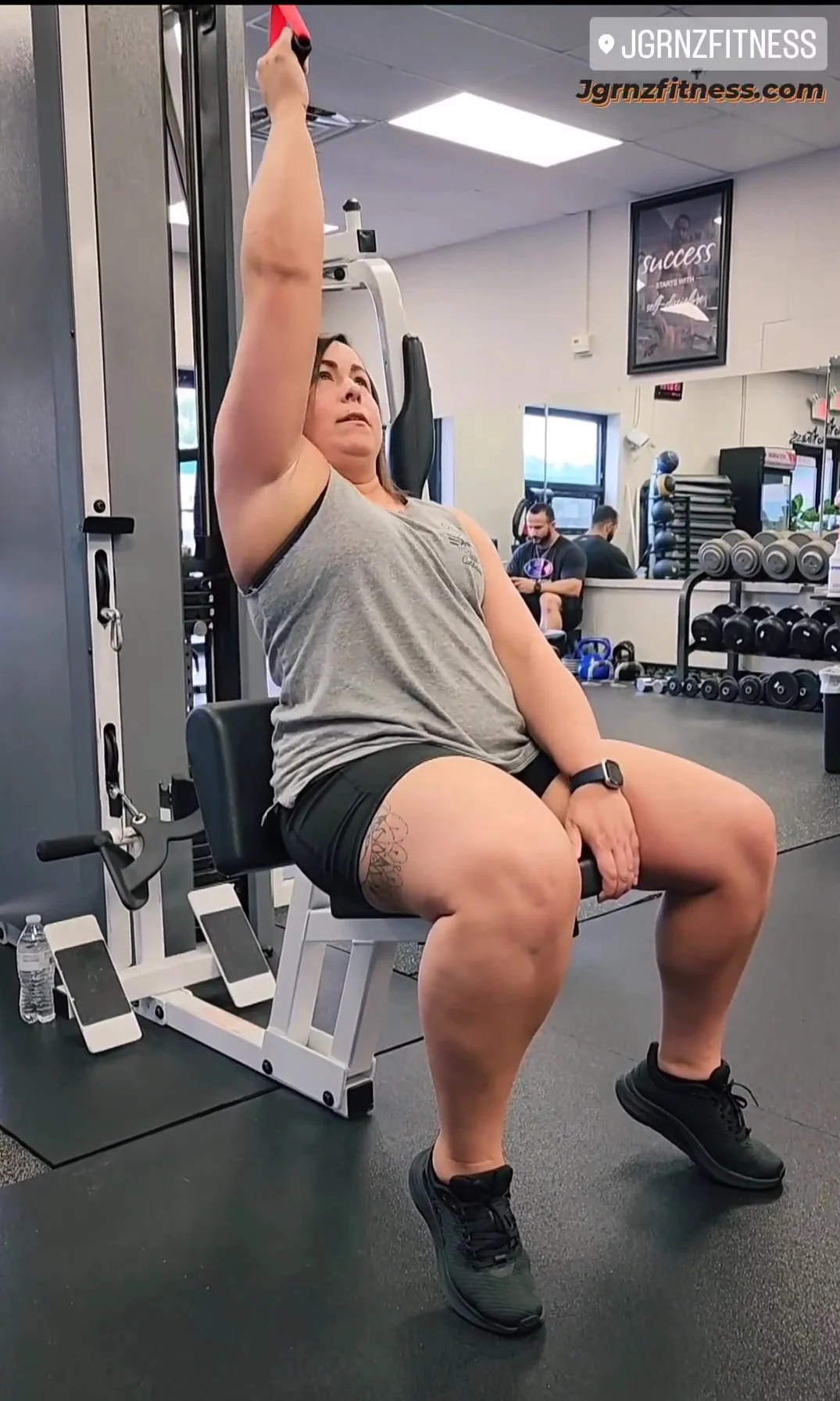 A woman sitting on a workout bench in a gym, raising her right arm with a weight in her hand, engaging in an exercise. She is wearing a gray sleeveless top, black shorts, and black athletic shoes, with a tattoo on her left thigh and a smartwatch on h