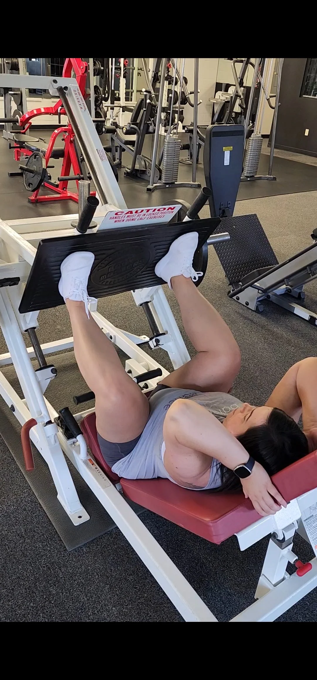 A woman lying on a workout bench in a gym, doing a leg press exercise with her feet on a footplate.