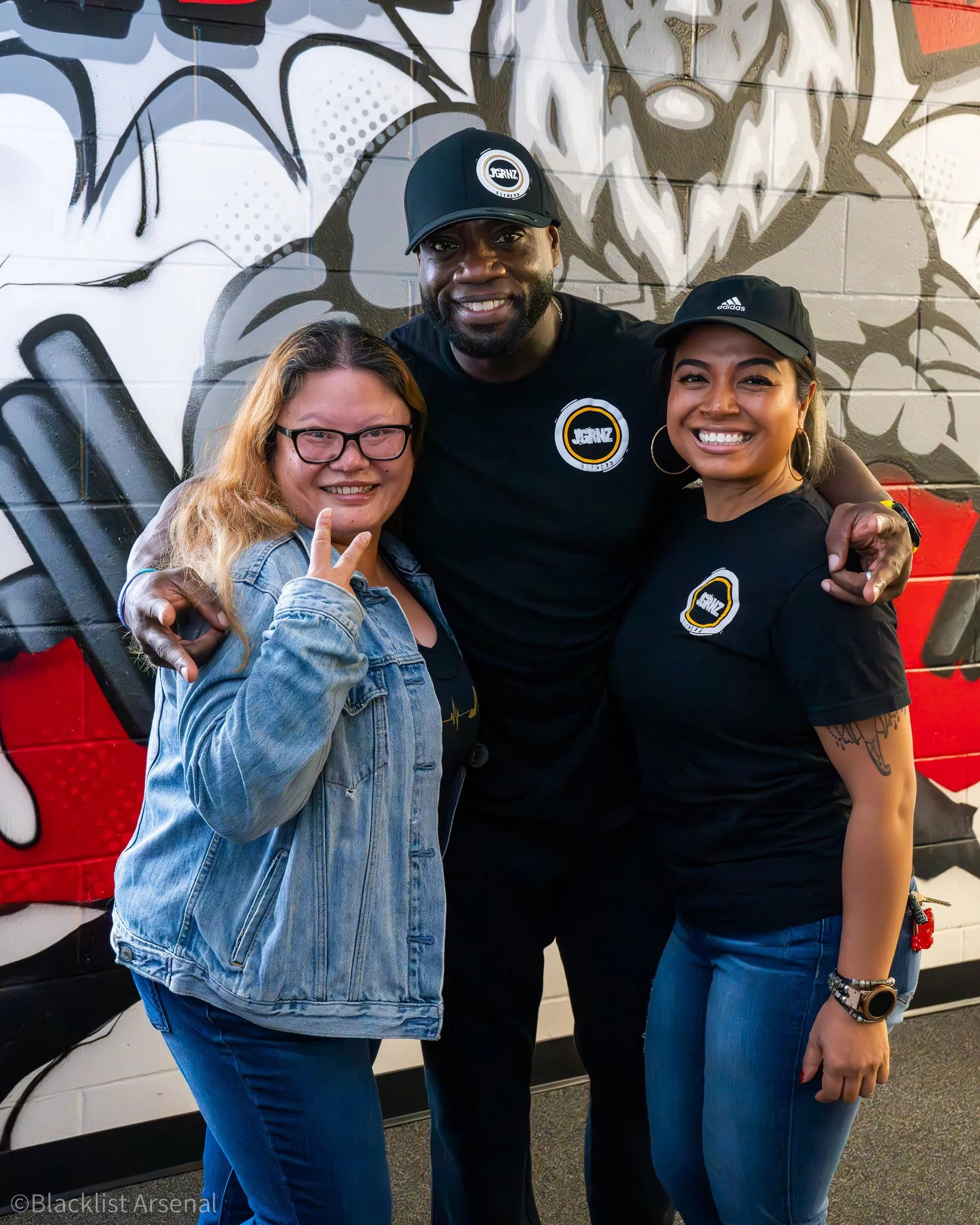 Three people smiling and posing in front of graffiti art wall, with two women and one man, all wearing branded clothing and hats.