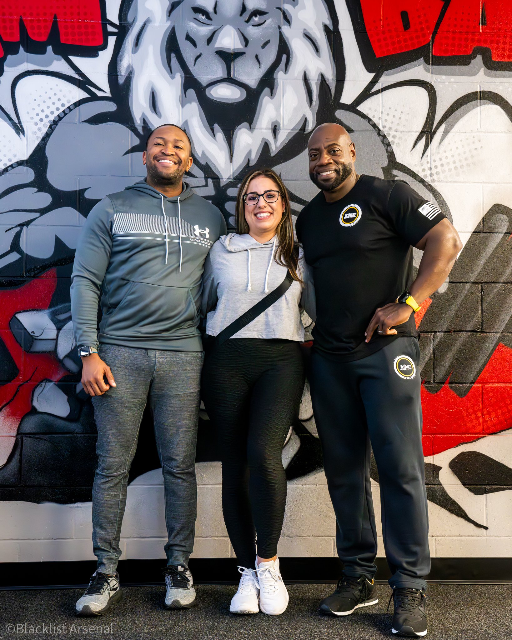 Three smiling people standing in front of a large graffiti mural of a lion on a wall, wearing athletic clothing.