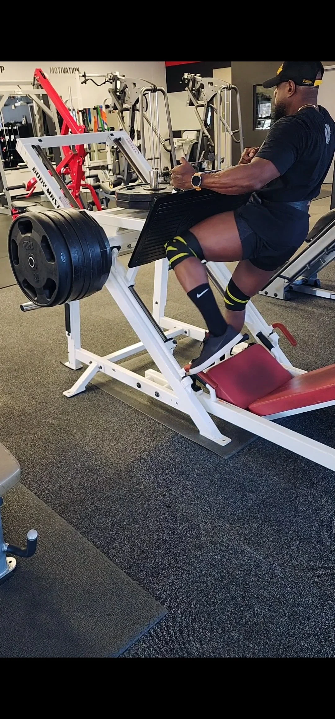 A man is performing a leg press exercise at a gym, wearing athletic clothing, including a black t-shirt, shorts, and knee-high socks with a Nike logo.