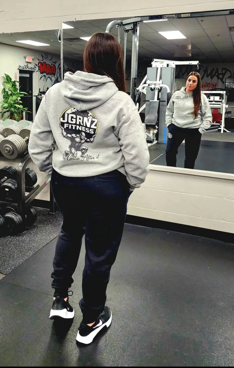 A woman with brown hair wearing a gray hoodie and black workout pants stands in front of a gym mirror, looking at her reflection. There are weightlifting equipment and dumbbells in the background.