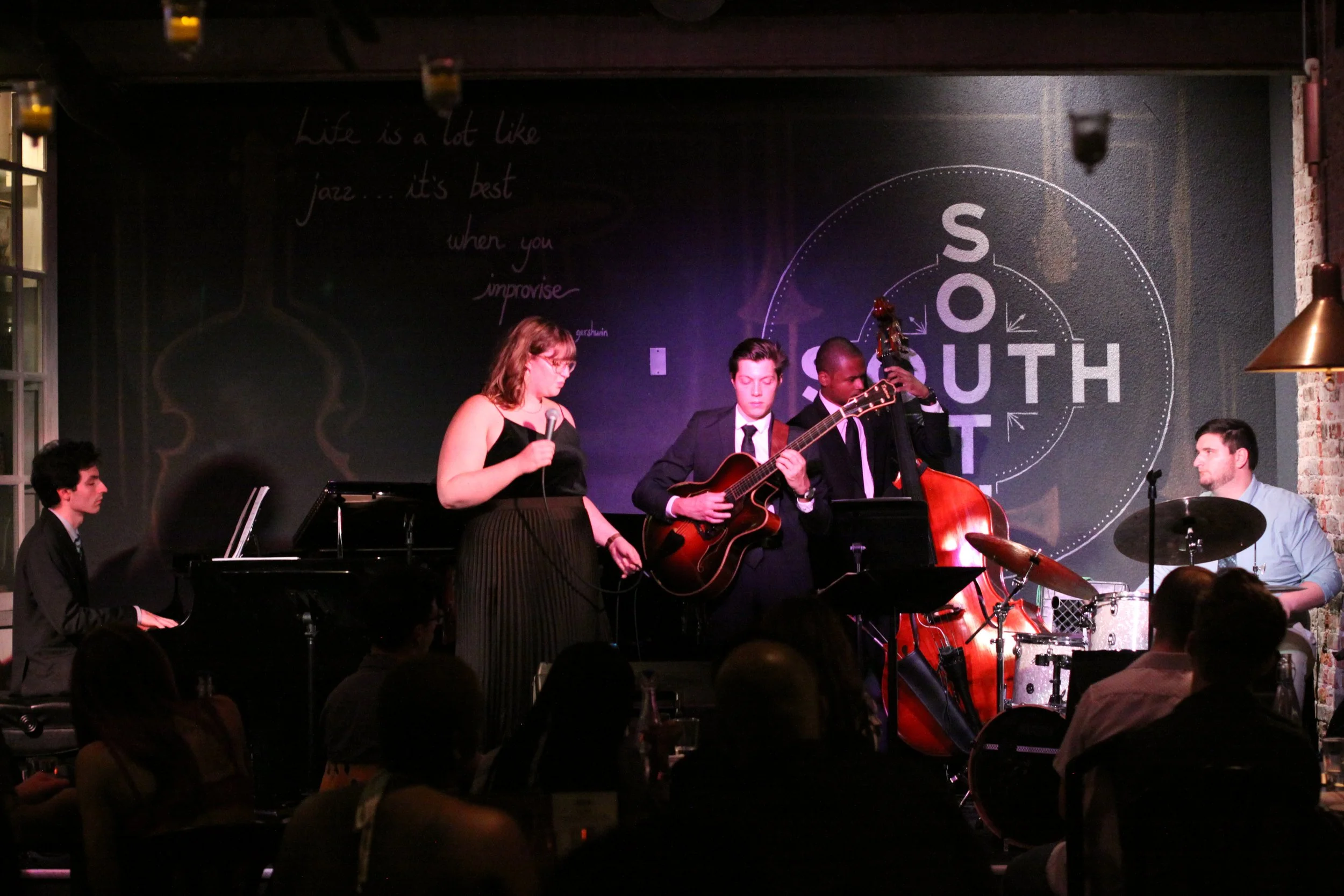 Jazz band performing live on stage with a pianist, a vocalist, a guitarist, a double bass player, and a drummer, at a venue with the words 'South' and 'Life is a lot like jazz' on the wall.