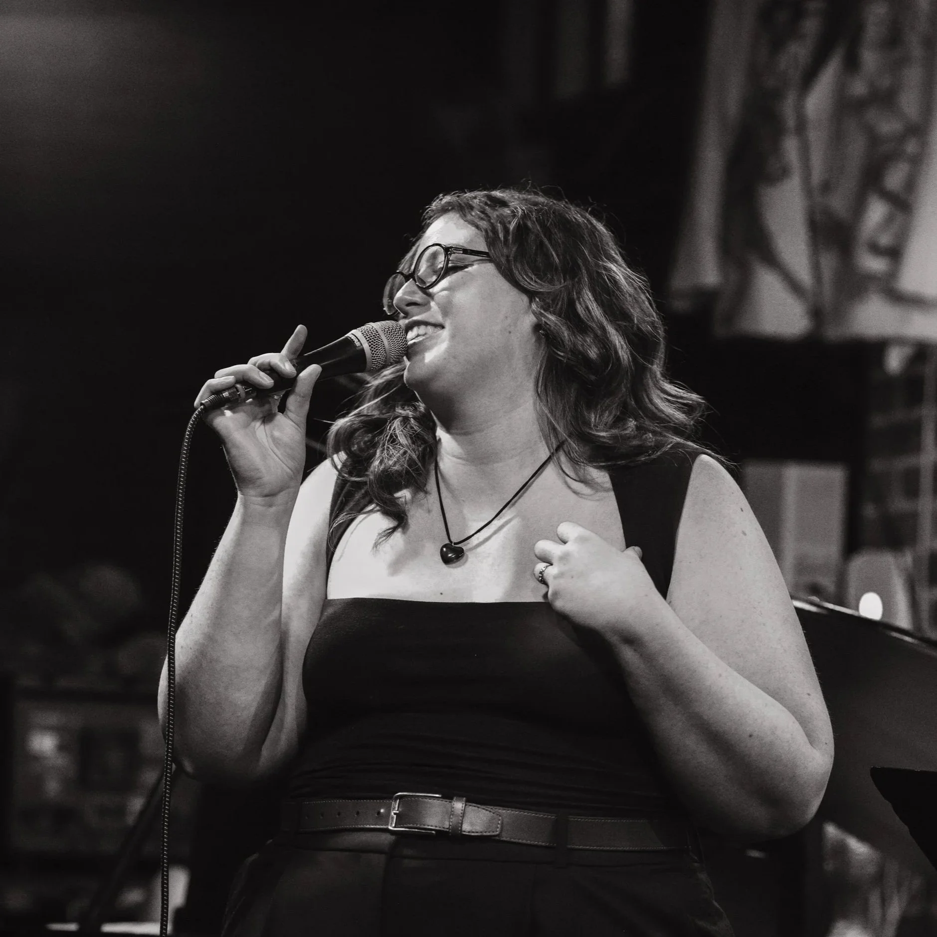 A woman singing into a microphone on stage, wearing glasses, a black sleeveless top, and a necklace with a heart pendant, smiling with her eyes closed.