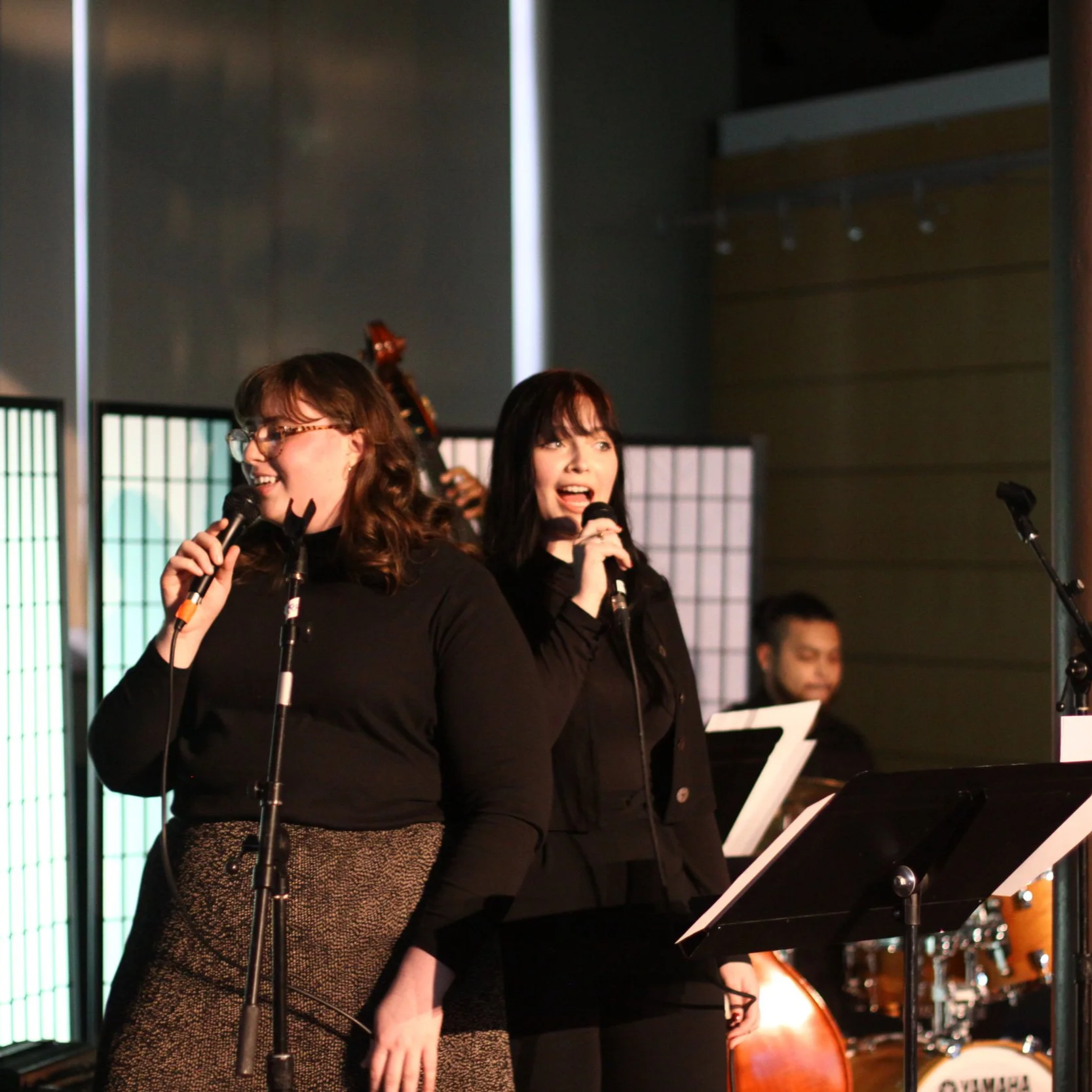 Two women singing into microphones on stage during a musical performance, with a band member and musical instruments in the background.