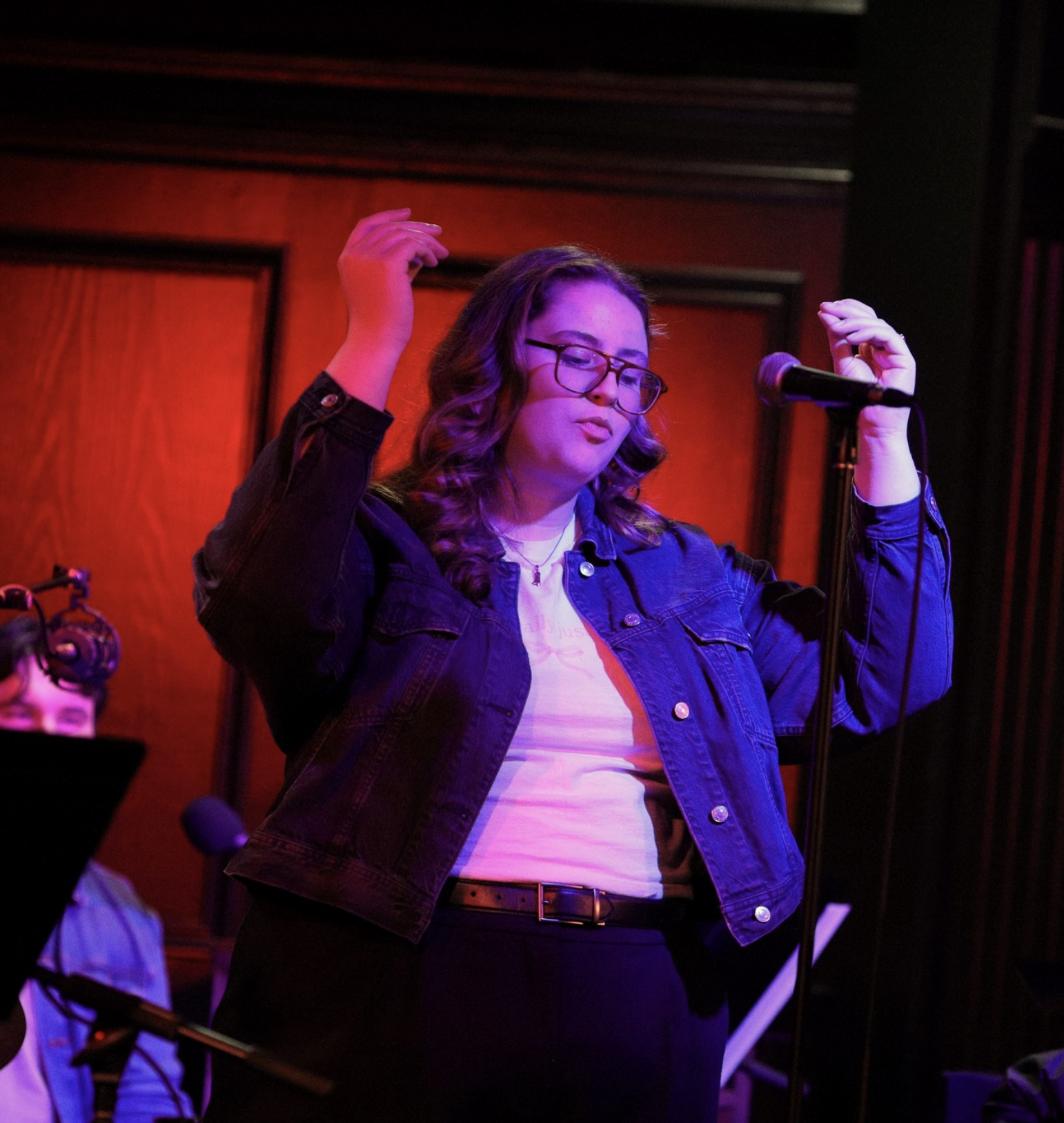 A woman with glasses, curly hair, wearing a denim jacket, standing in front of a microphone, with a red background, appearing to sing or perform.
