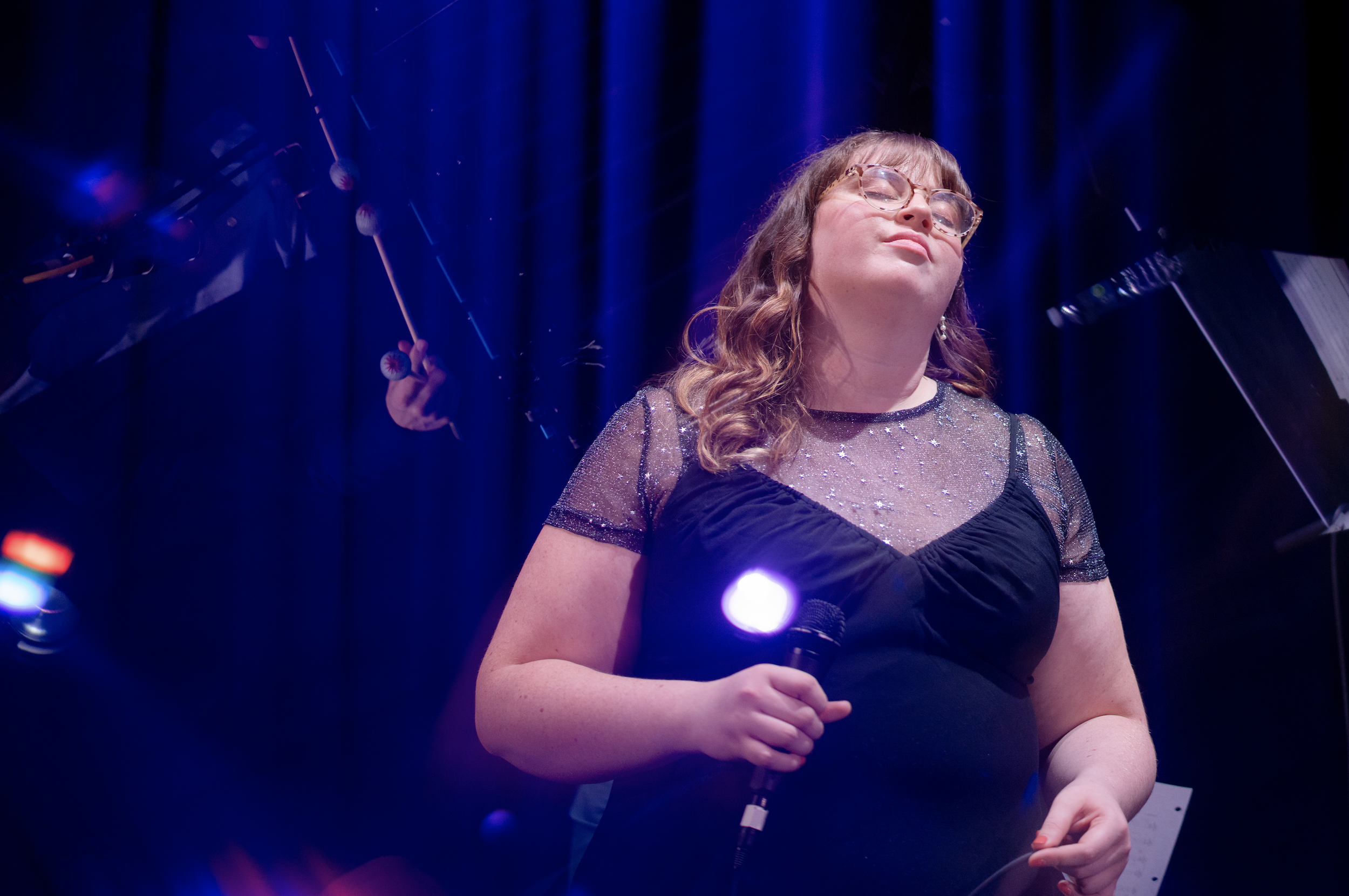 A young woman with glasses, long wavy red hair, wearing a black dress with a sheer, sparkly top, stands on stage holding a microphone with a purple light. She has a confident expression, with her eyes closed and head slightly tilted back. The backgro