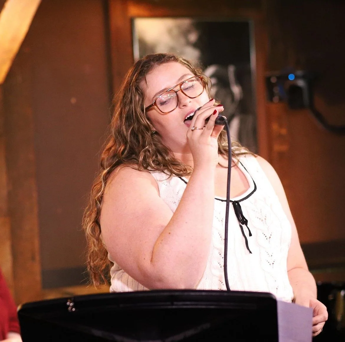A woman with curly hair and glasses sings into a microphone, eyes closed, in a cozy indoor setting.
